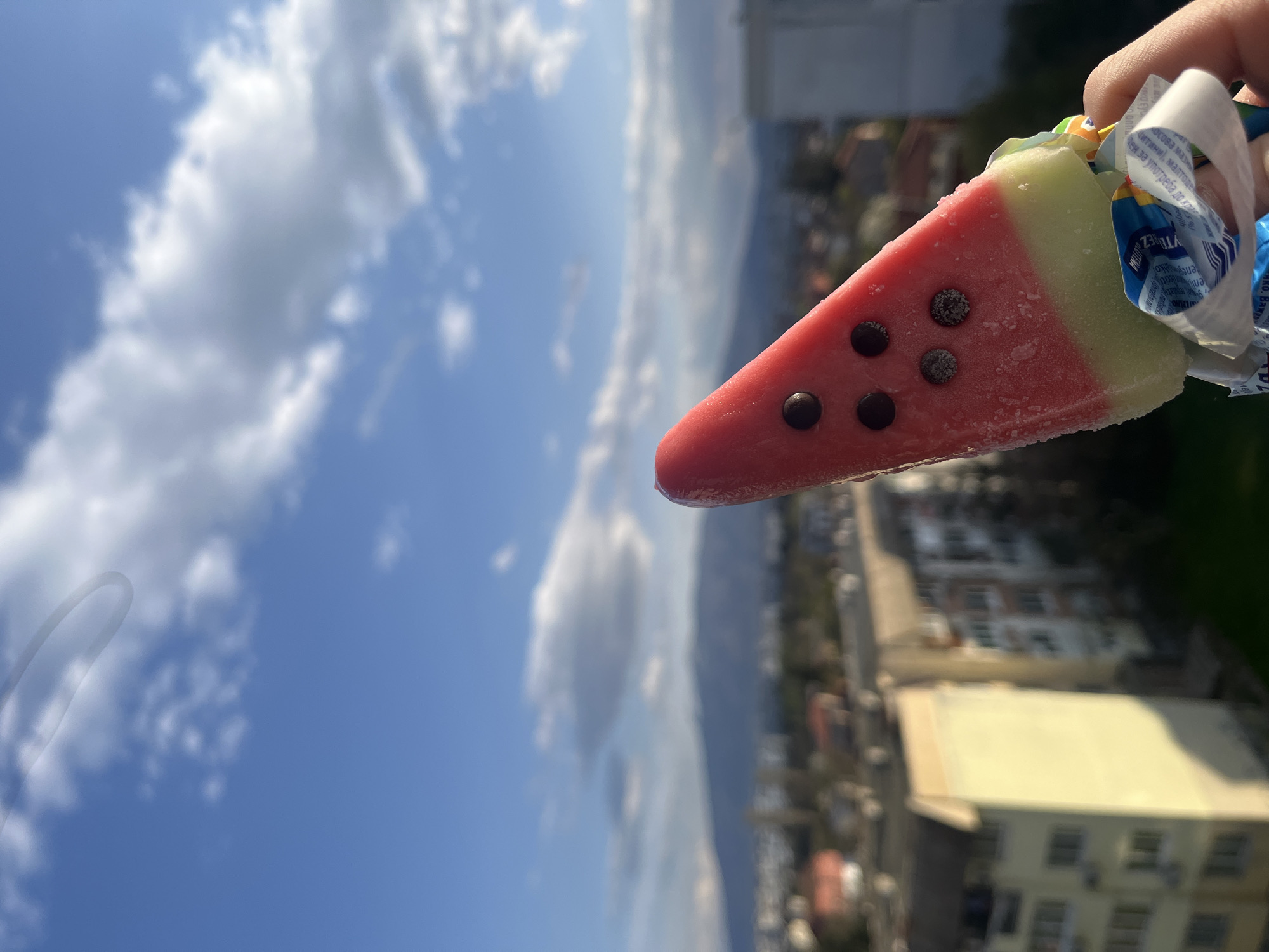 The photograph depicts a watermelon-flavored popsicle held against a backdrop of a cityscape and a cloudy, blue sky. The popsicle is shaped like a slice of watermelon, with a red section containing chocolate chips to mimic seeds, and a green rind. The wrapper is partially peeled down, revealing a small portion of the stick. The cityscape in the background appears to be residential, with buildings of varying heights and colors. The scene is likely in Sofia, Bulgaria, though the presence of landmarks is insufficient to establish an exact location. No people are visible other than the hand holding the popsicle. The environment suggests a clear, sunny day.