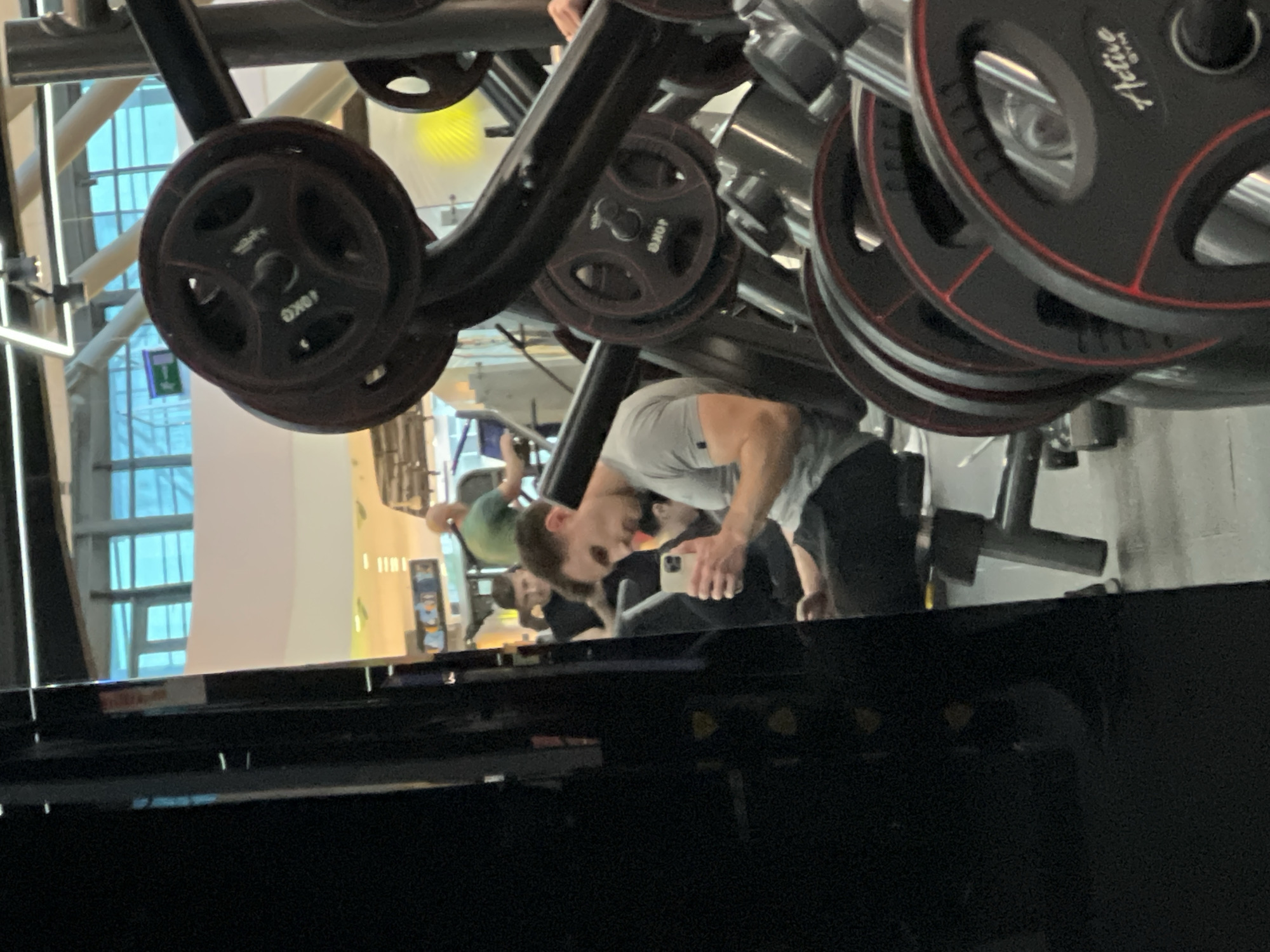 The image shows an indoor gym setting with gym equipment, including weight plates. A man in a light-colored shirt and dark pants is looking at a phone. Another person in a green shirt is visible in the background. The gym has large windows. This appears to be a reflection in the surface in the lower portion of the frame. The location is implied to be Burgas, Bulgaria, though visual verification is not possible from the content of the image.