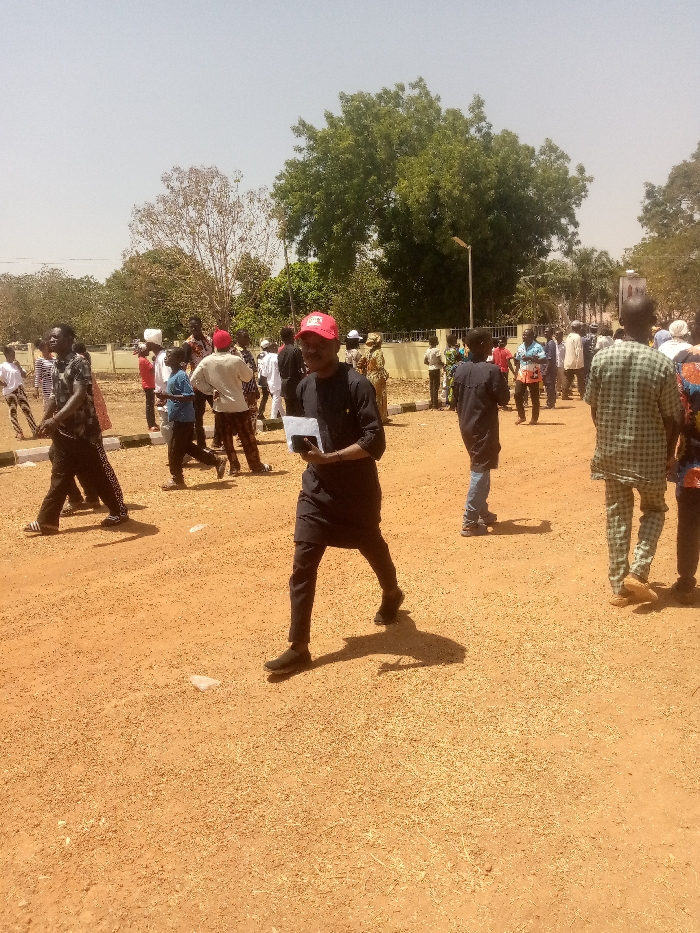 The image depicts an outdoor scene in Kagoma, Nigeria, under bright, clear daylight. The ground is unpaved, consisting of reddish-brown earth with scattered small gravel and some white debris. In the background, green trees are visible, along with a low, light-colored wall or fence. A street lamp stands behind this wall.
Approximately 20-30 individuals are present, predominantly males, dispersed across the scene. In the foreground, a male subject walks from left to right, looking forward. He wears a dark long-sleeved tunic, dark trousers, and dark shoes. A red baseball cap is on his head. He holds a small, white rectangular object in his left hand and a dark rectangular object, consistent with a smartphone, in his right hand. His shadow is cast distinctly to his right.
To the left and slightly behind him, another male walks right, wearing a patterned dark shirt, dark trousers with white stripes, and sandals. His shadow is also visible. To the right, another male subject is seen from the back, wearing a light green and white patterned garment with matching trousers.
Other individuals are scattered throughout the mid-ground and background, wearing various casual and traditional clothing. Some are standing, others are walking. No specific interactions are clearly discernible, but the individuals appear to be moving or gathered in an open area. Shadows are harsh and defined, indicating direct overhead sunlight.