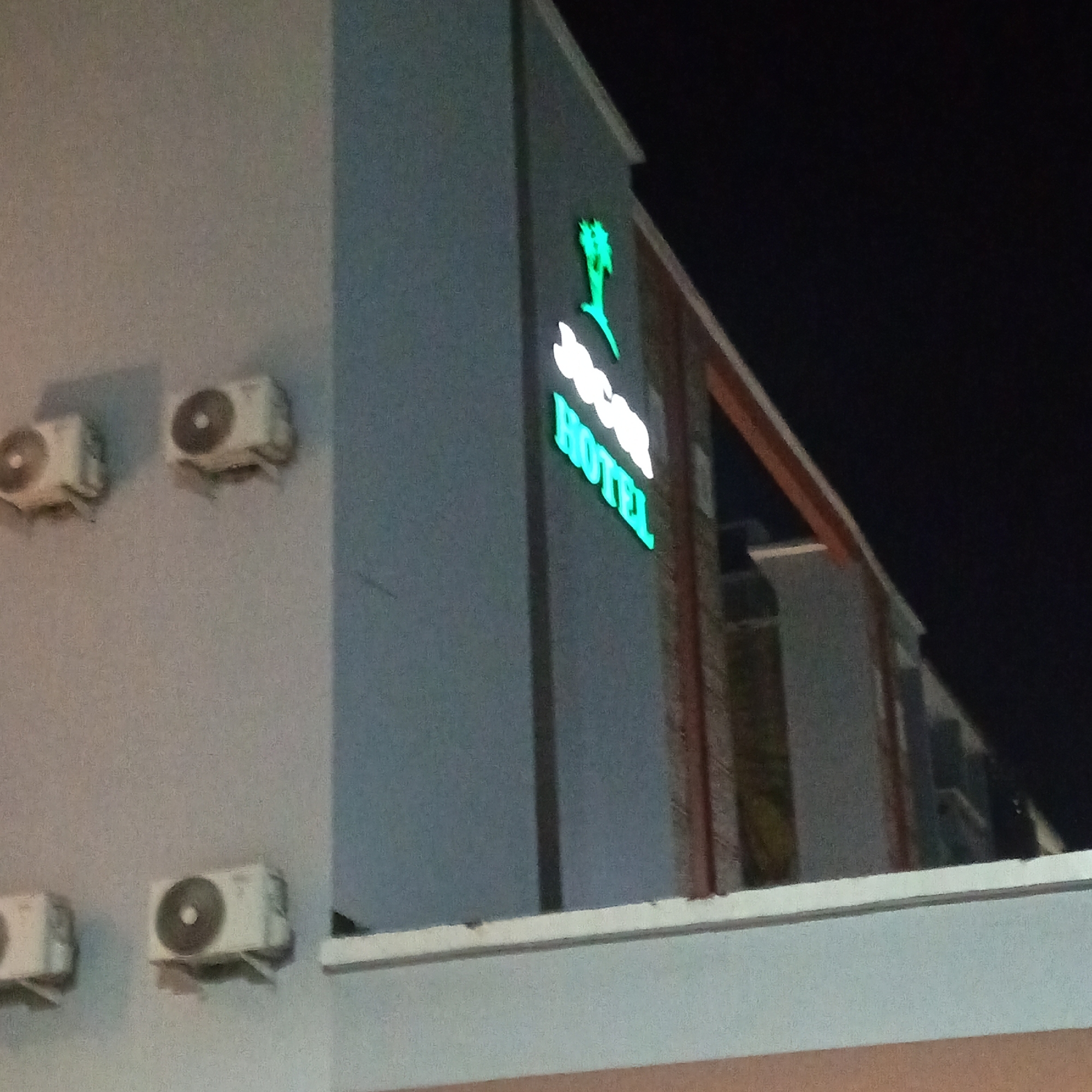 The image shows the exterior of the "Sears Hotel" in Ibadan, Nigeria, at night. A neon sign with the hotel's name and a palm tree graphic is visible, illuminated in green and white. The building is a light gray color and has several outdoor air conditioning units mounted on its exterior wall. The sky is dark, indicating it is nighttime. The image is taken from a low angle, looking up at the hotel.