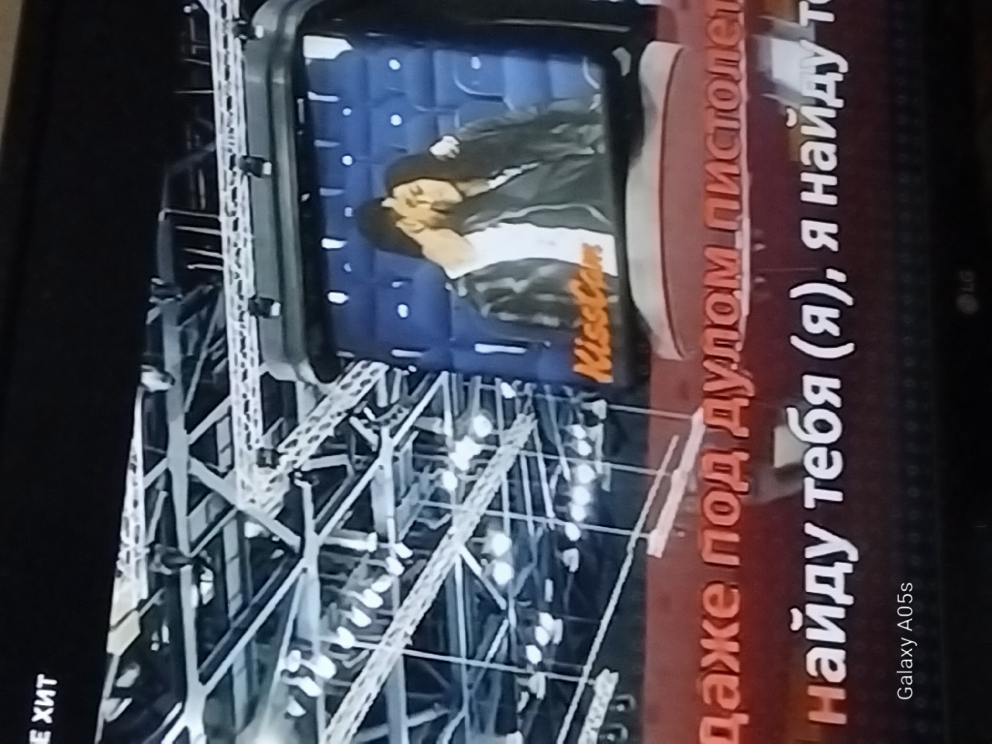 The image displays a television screen showing a "KissCam" segment. In the center of the screen, a couple is shown embracing. The background of the television display is a blue stadium seating area. Below the couple, text in Russian is visible, including the words "даже под дулом пистолета" which translates to "even under the muzzle of a pistol," and "найду тебя (я), я найду т" which translates to "I will find you (I), I will find t". In the lower left corner of the screen, the text "Galaxy A05s" is displayed, indicating the device used to capture the image. The overall environment appears to be indoors, possibly a stadium or concert hall, suggested by the lighting rigs and structural elements visible in the background behind the television. The specific location in the city of Druzhba, Russia is not discernible from the provided image.