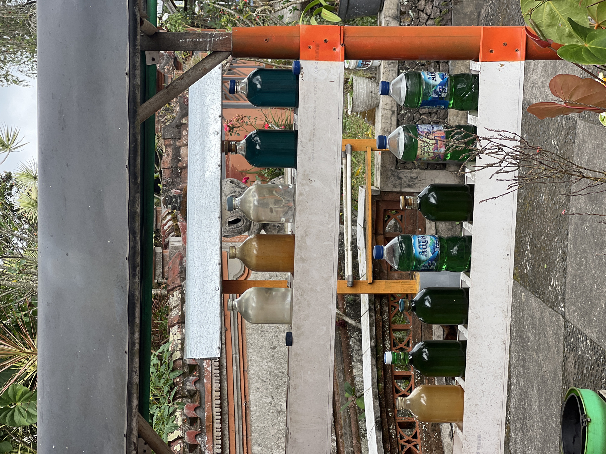 In Rendang, Indonesia, a roadside stall displays various liquids in bottles, likely for sale. The stall has two tiers, each holding several bottles. The top tier features three glass bottles containing amber-colored liquid and one clear glass bottle filled with a greyish liquid. To the right, two darker glass bottles hold a deep green liquid. The bottom tier showcases four plastic bottles of green liquid, one of which is clearly labeled "AQUA" and contains clear water. Adjacent to the AQUA bottle is another identical bottle, also with green liquid. The stall is constructed from metal and wood, supported by orange-colored poles. The background reveals a typical Indonesian setting with lush greenery, red-tiled roofs, and stone walls. The ground is paved with grey stones. The scene is devoid of people.