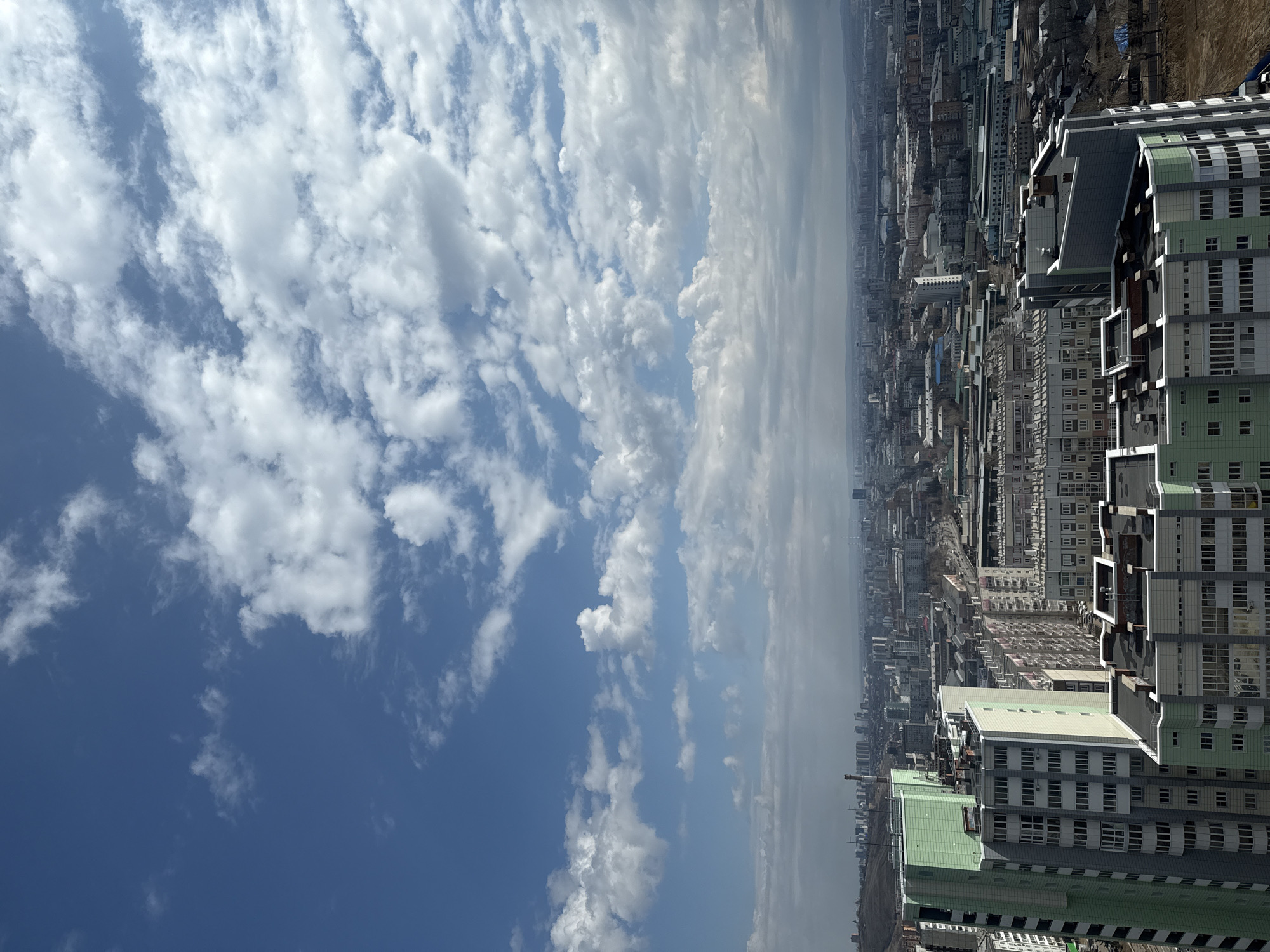 The image depicts an elevated view of Krasnoyarsk, Russia, on a partly cloudy day. The sky is predominantly blue with scattered cumulus clouds. Below, the cityscape extends to the horizon, showcasing dense urban development with numerous buildings and structures. In the foreground, a cluster of tall apartment buildings with light green accents is visible. The city features a mix of building heights and architectural styles, from older, smaller structures to modern high-rises. The air appears slightly hazy, especially in the distance. The general environment suggests an urban setting with typical infrastructure. No people are visible.