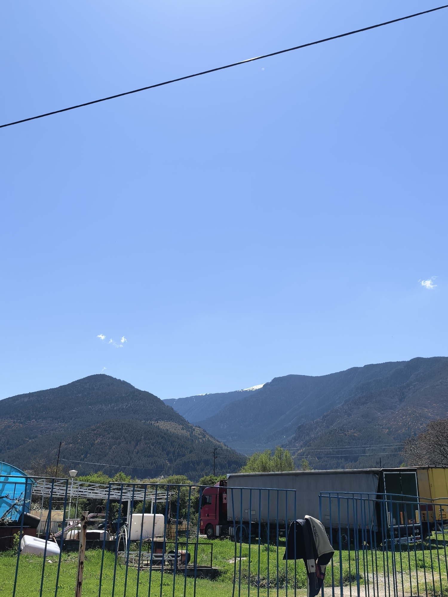 The image shows a landscape with mountains in the background under a clear blue sky. In the foreground, there's a fence with what appears to be a grassy area. Behind the fence are various objects including a truck and some miscellaneous items. A power line is visible across the top of the image. The location is described as "Unknown, Unknown".
