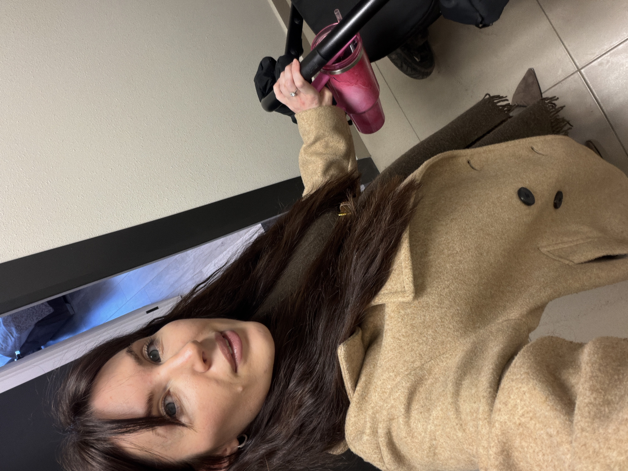 The image depicts a female individual taking a selfie indoors, positioned in what appears to be a residential entryway or hallway. The female, with long dark brown hair and bangs, light skin, and blue eyes, looks directly at the camera. She wears a light tan or beige wool-blend coat with two dark buttons visible, and a dark olive-green fringed scarf is partially visible beneath the coat. A silver ring with a clear stone is present on her left ring finger, and a small silver hoop earring is in her left ear. Her right hand grips the black handle of a black stroller or pram, which also holds a pink insulated tumbler with a straw. In the background, a dark doorframe opens to an adjacent room where a bed with light-colored bedding is partially visible. The walls are a textured off-white, and the floor is covered with light-colored square tiles. The specific location within the city of Perm, Russia, cannot be determined from the visual evidence in the image.