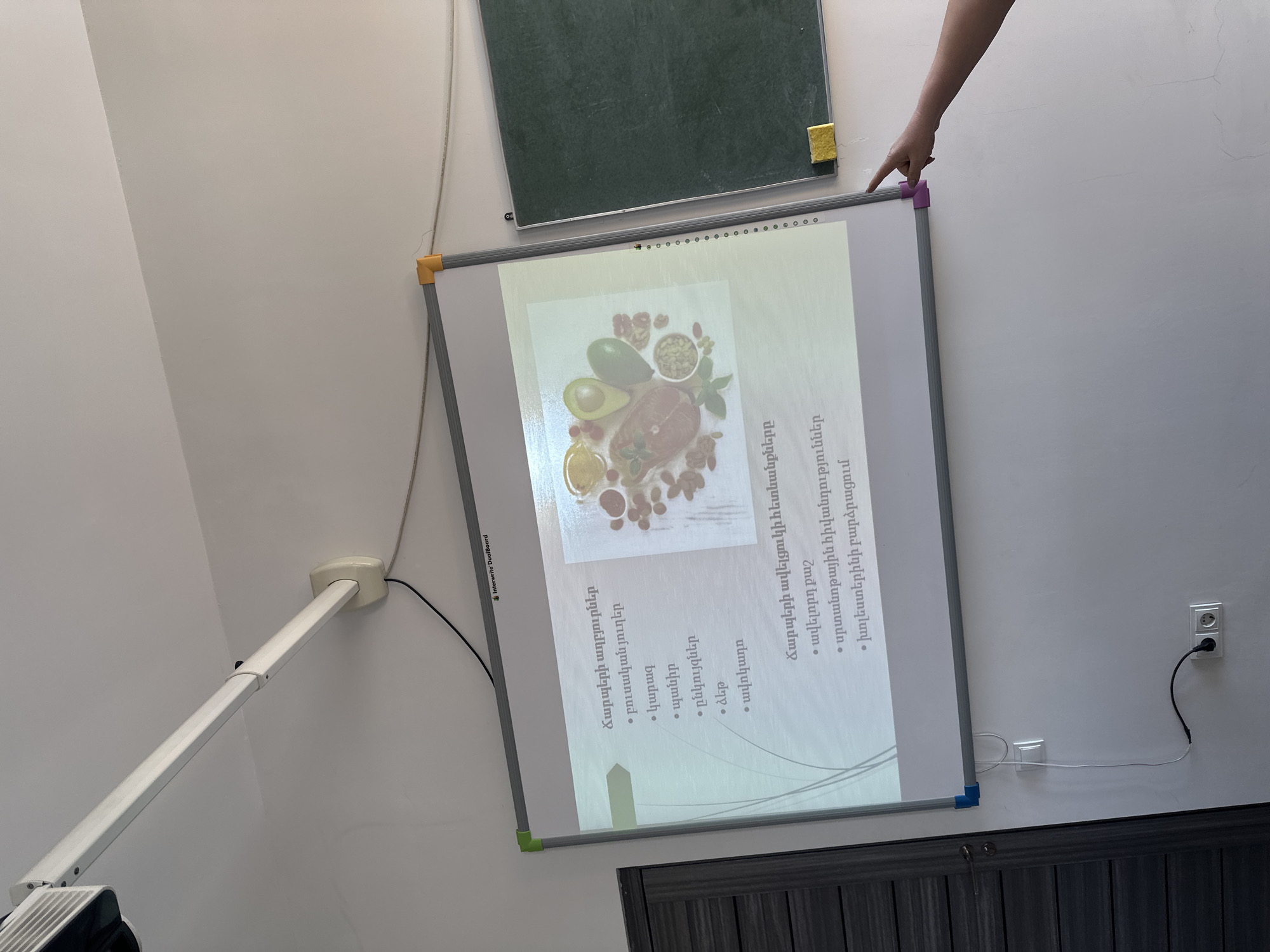 Here is a matter-of-fact description of the scene:
An image shows a presentation board mounted on a wall. The presentation screen displays an image of food items like avocados, nuts, and other produce. Text in Armenian is visible on the screen, discussing sources of fat and the consequences of excessive fat intake. A hand points at the top corner of the presentation board. Above the presentation board is a green chalkboard, and to the right is an electrical outlet. There's a projector stand with a visible portion of a white projector attached. The bottom of the frame shows a dark colored wainscotting on the lower portion of the wall.