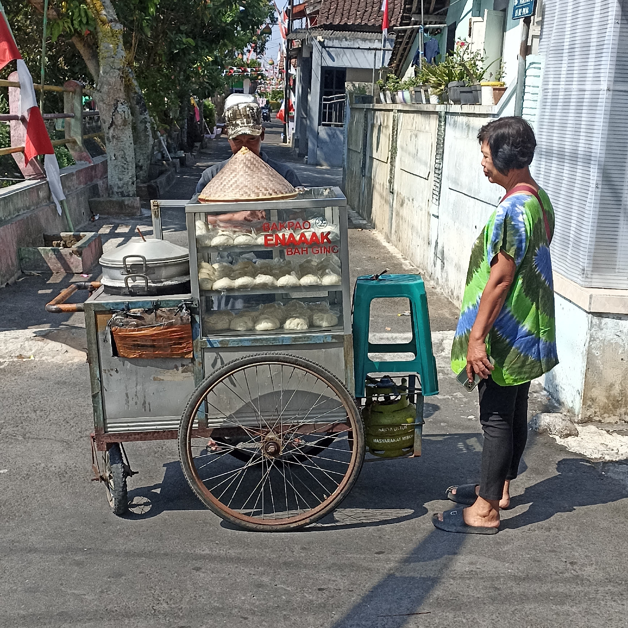 In Magelang, Indonesia, on a sunlit street, a food cart vendor, a person wearing a hat and traditional conical bamboo hat, stands behind a glass-fronted display filled with steamed buns. The cart is equipped with a steaming pot, a green gas cylinder, and a teal plastic stool. A woman, wearing a vibrant green and blue tie-dye shirt and black pants, stands facing the cart, holding a smartphone. The street is paved with asphalt and lined with buildings and some greenery. Decorative flags and banners are visible in the background, suggesting a local celebration or event. The scene captures a moment of commerce and daily life in the city.