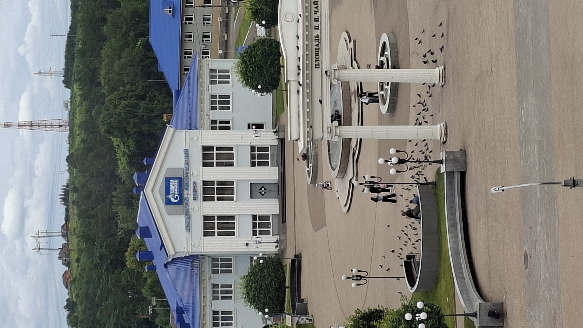 The image captures a view of the central square in Chaikovsky, Russia. In the foreground, a paved plaza is dotted with pigeons. Several people are scattered throughout the plaza, some sitting on a curved bench, others standing near a lamppost, and a child is riding a bicycle. Two multi-tiered fountains are visible, one with water flowing.
Dominating the mid-ground is a light-colored building with a prominent blue roof, featuring the Gazprom logo and text that appears to read "Культурно-спортивный центр" (Cultural and Sports Center). The building has large, multi-paned windows and an imposing central entrance. To the right of this building, another building with a similar blue roof is partially visible.
The background consists of a dense green forest on a hillside, with several tall industrial towers and power lines silhouetted against a cloudy sky. The overall scene depicts a public space in a Russian city, with elements of architecture, nature, and human activity.