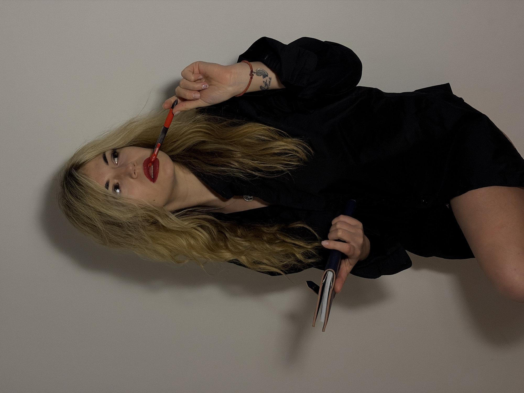 A woman with blonde hair and red lipstick is lying on her side against a light gray wall. She is wearing a black top. She holds a pen in her mouth and another pen and notebook in her right hand. A tattoo is visible on her left wrist. The location and city of Budva, Montenegro, are not evident from the image.