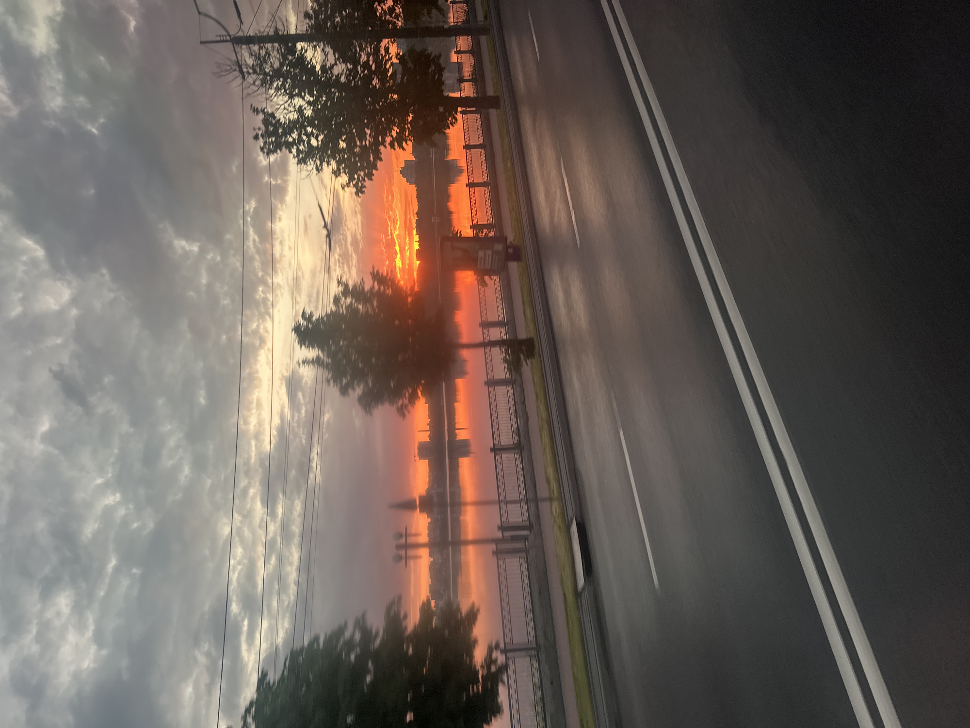 The image captures a sunrise over the Ternopil Reservoir in Ternopil, Ukraine. The sky is filled with dramatic clouds, illuminated by the vibrant orange and red hues of the rising sun. The sun is partially obscured by clouds, casting a fiery glow on the water's surface. Along the reservoir's edge, a promenade is visible, featuring a decorative metal fence and trees lining the pathway. Beyond the promenade, city buildings can be seen in the distance, silhouetted against the bright sky. In the foreground, a road with white lane markings stretches across the frame, suggesting the perspective is from a moving vehicle. The road appears wet, reflecting the diffused light of the early morning. There are no people or animals clearly visible in the frame. The overall scene depicts a serene and visually striking urban landscape at dawn.
