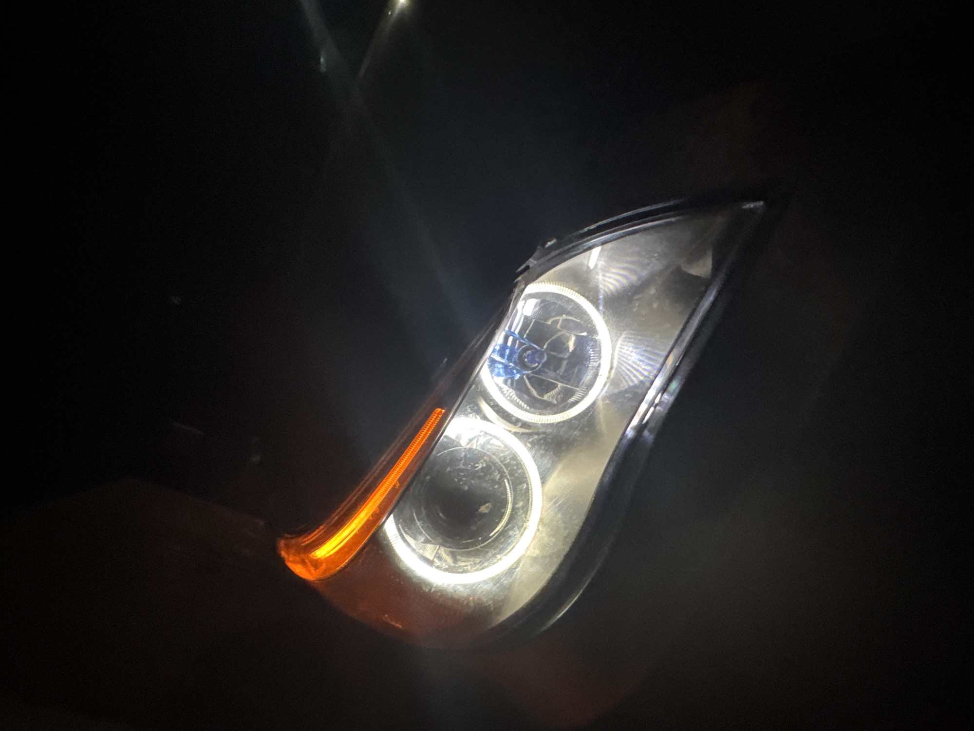 The image shows a close-up view of a car headlight at night. The headlight is illuminated, featuring two circular lamps with a bright halo effect. The turn signal section is orange. The surroundings are dark and indistinct, suggesting a low-light environment. The location (Sarnevo, Bulgaria) is not discernible from the image.