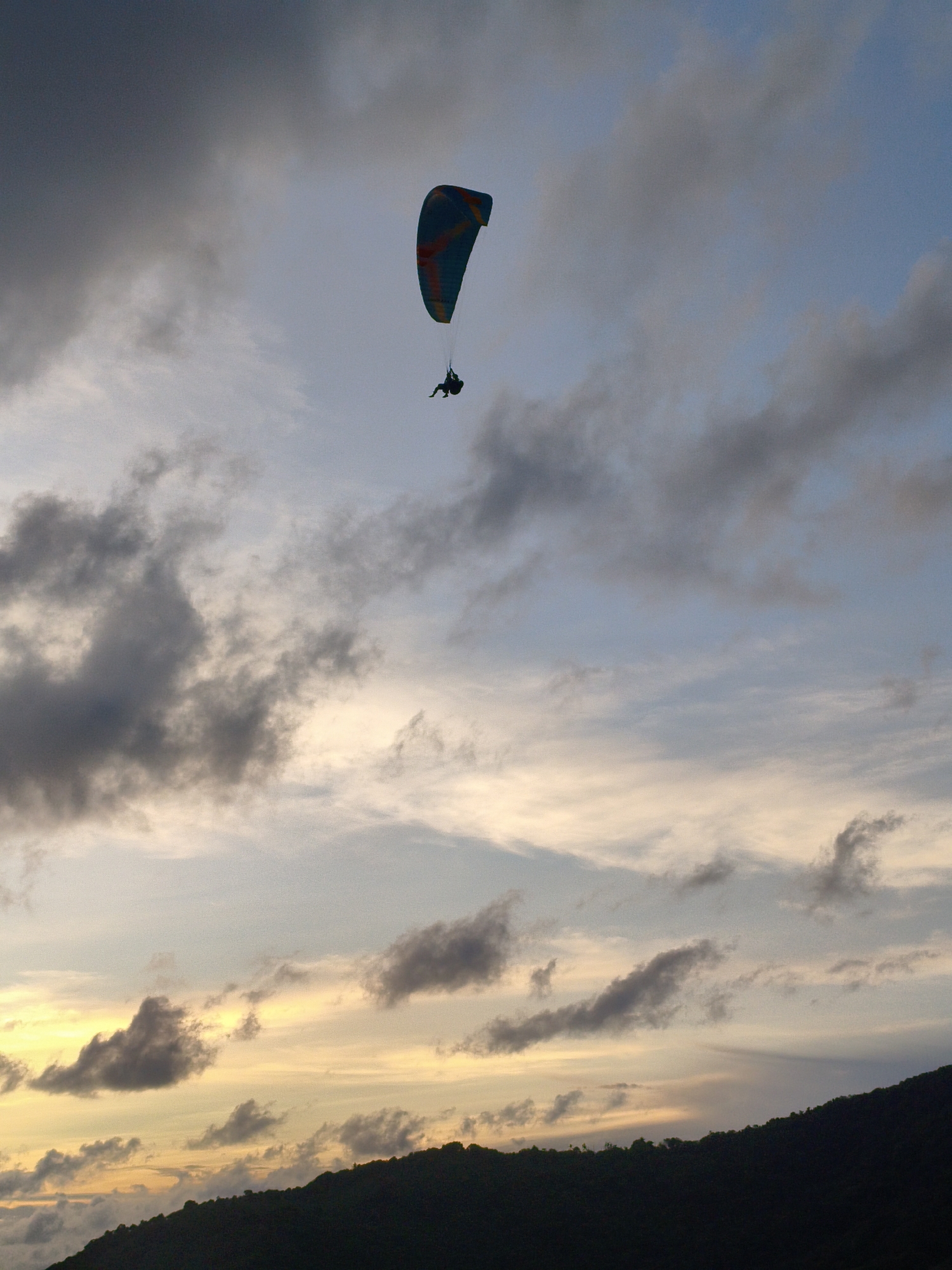 A single paraglider is captured mid-flight against a dramatic twilight sky. The paraglider's canopy is a vibrant mix of blue and orange, contrasting with the surrounding clouds. A person is suspended beneath the canopy, their silhouette dark against the lighter sky. The background is filled with a gradient of colors, from the deep blues and grays of approaching night to the soft yellows and oranges of the setting sun near the horizon. A dark, silhouetted mountain range forms the bottom edge of the image, indicating a landscape setting.
