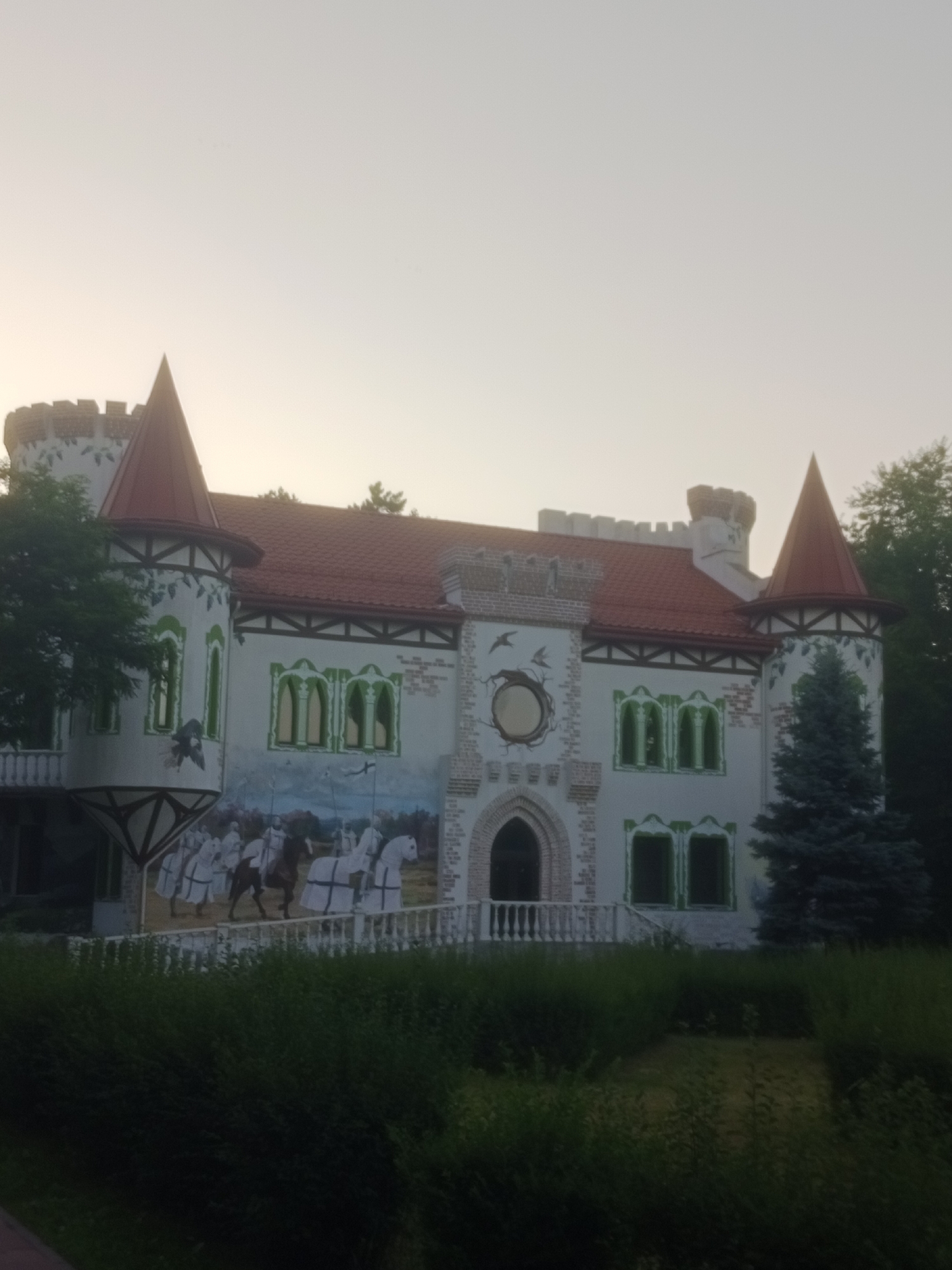 The image shows a fairy tale castle-like building with a white facade, red tiled roofs, and green accents around the windows. The castle has multiple towers, battlements, and decorative elements. A mural depicting knights on horseback is painted on the side of the building. The castle is surrounded by greenery, including trees and bushes. The sky is pale and overcast, suggesting it is either early morning or late evening. The building appears to be a tourist attraction or a themed establishment. This structure is known as the "Fairy Tale Castle" located in the city of Horishni Plavni, Ukraine.