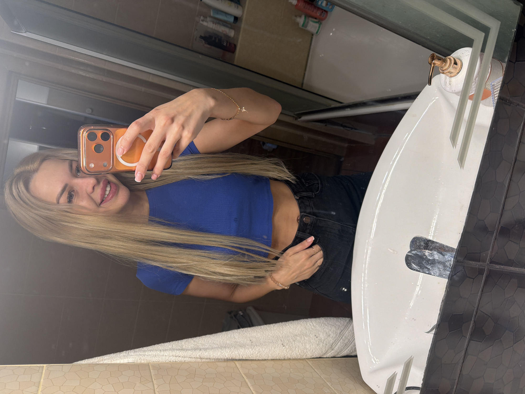 A young woman with blonde hair is taking a selfie in a bathroom mirror. She is wearing a blue crop top and black jeans. She is holding an orange phone with a rose gold case. The bathroom appears to be small and tiled. There is a sink in the foreground, and a bathtub and shelves with toiletries are visible in the reflection. The scene is likely indoors in a residential setting, possibly in Kyiv, Ukraine, given the provided context. The lighting is artificial, suggesting an indoor environment.