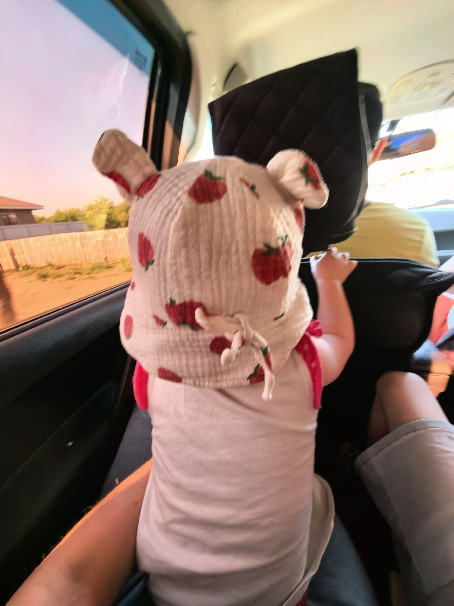 A blurry, slightly tilted, eye-level shot from inside a moving car shows a toddler wearing a strawberry-print hat and white onesie. The child is facing forward, arms up near the back of the front passenger seat. A person's arm and leg are visible in the foreground, indicating they are holding the child. Outside the car window, a light-colored house and a wooden fence are visible against a pink sky, suggesting sunset or sunrise. The location is Sterlitamak, Russia.