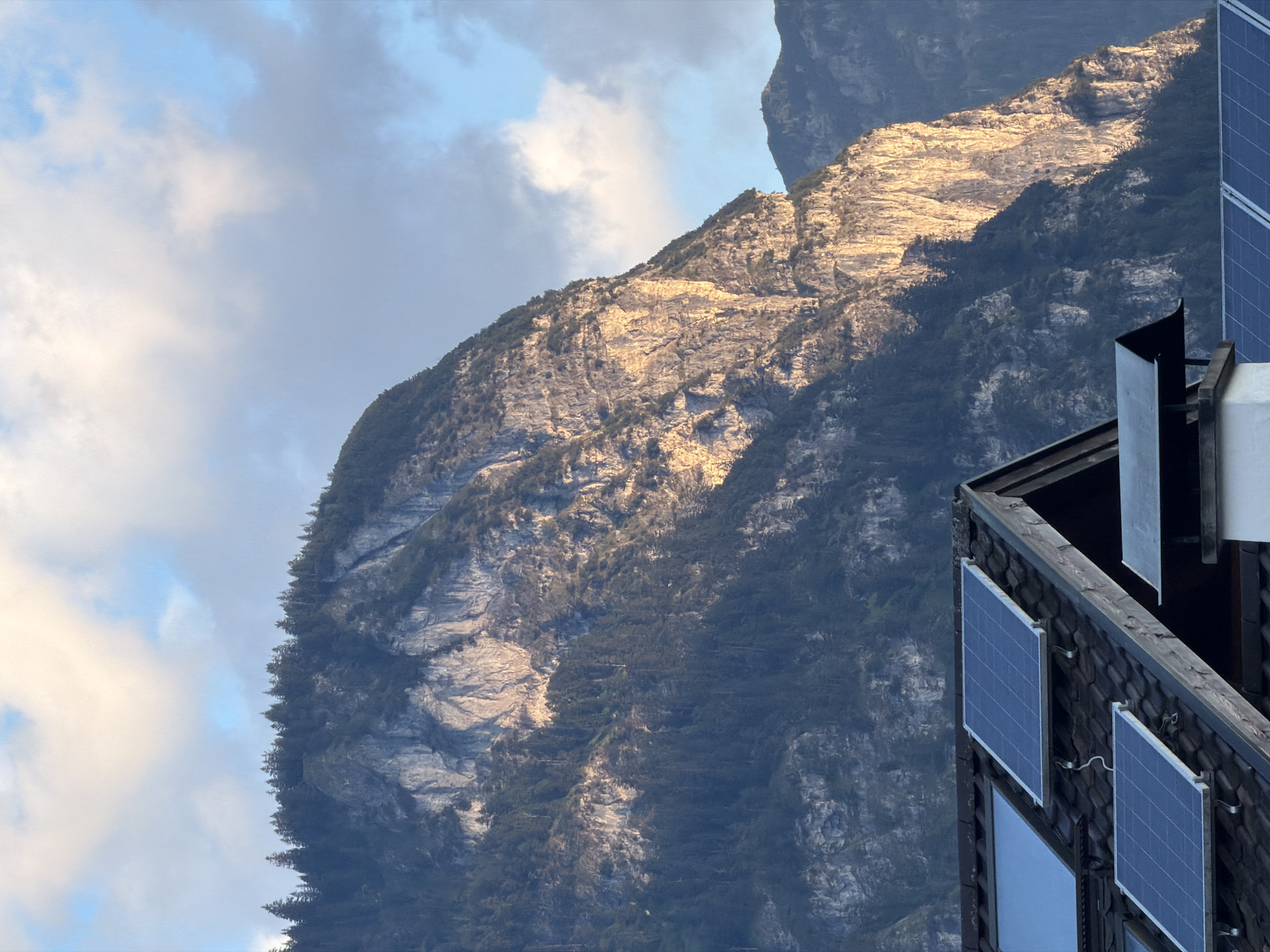 A close-up, low-angle shot captures a mountain range in Garmisch-Partenkirchen, Germany, with a residential rooftop in the foreground. The sun, low in the sky, casts a warm glow on the rugged, rocky mountain face, highlighting its texture and the dark green coniferous forest clinging to its slopes. The sky above is a mix of blue with scattered, soft white clouds. The foreground features a dark tiled roof with several solar panels installed. Part of a chimney is also visible. The scene suggests late afternoon or early morning light.