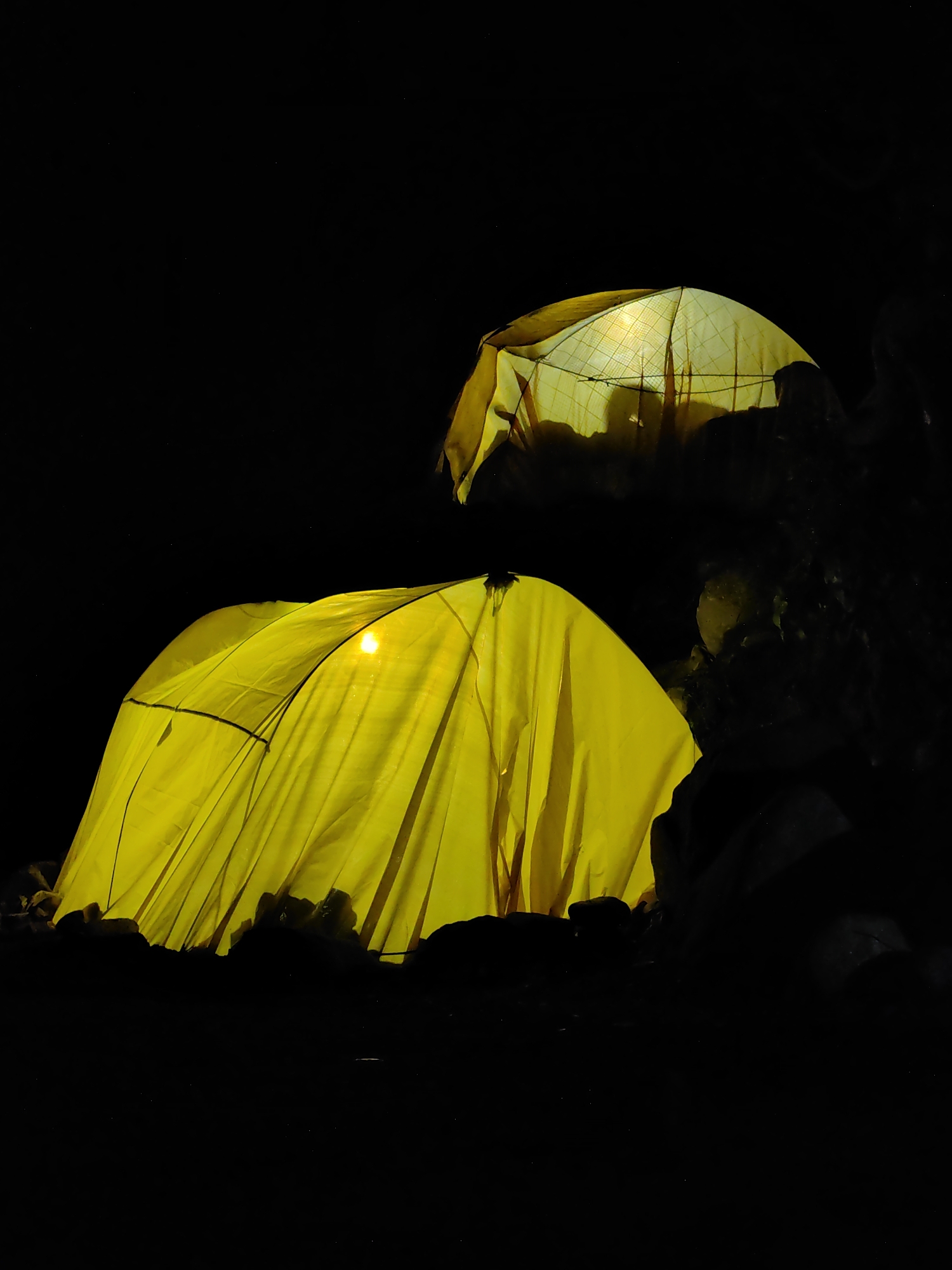 The image depicts two illuminated yellow tents set up in a dark, rocky environment. The tent in the foreground is larger and more prominently lit, with a bright light source visible inside, casting shadows and highlighting the fabric's texture. The second tent, positioned behind and slightly to the right of the first, is also lit from within, revealing a patterned interior and a silhouette of at least one person. The surrounding area is dark, with indistinct rocks and foliage silhouetted against the illuminated tents. The overall scene suggests a camping or outdoor setting at night. The location is identified as Palchan, India.