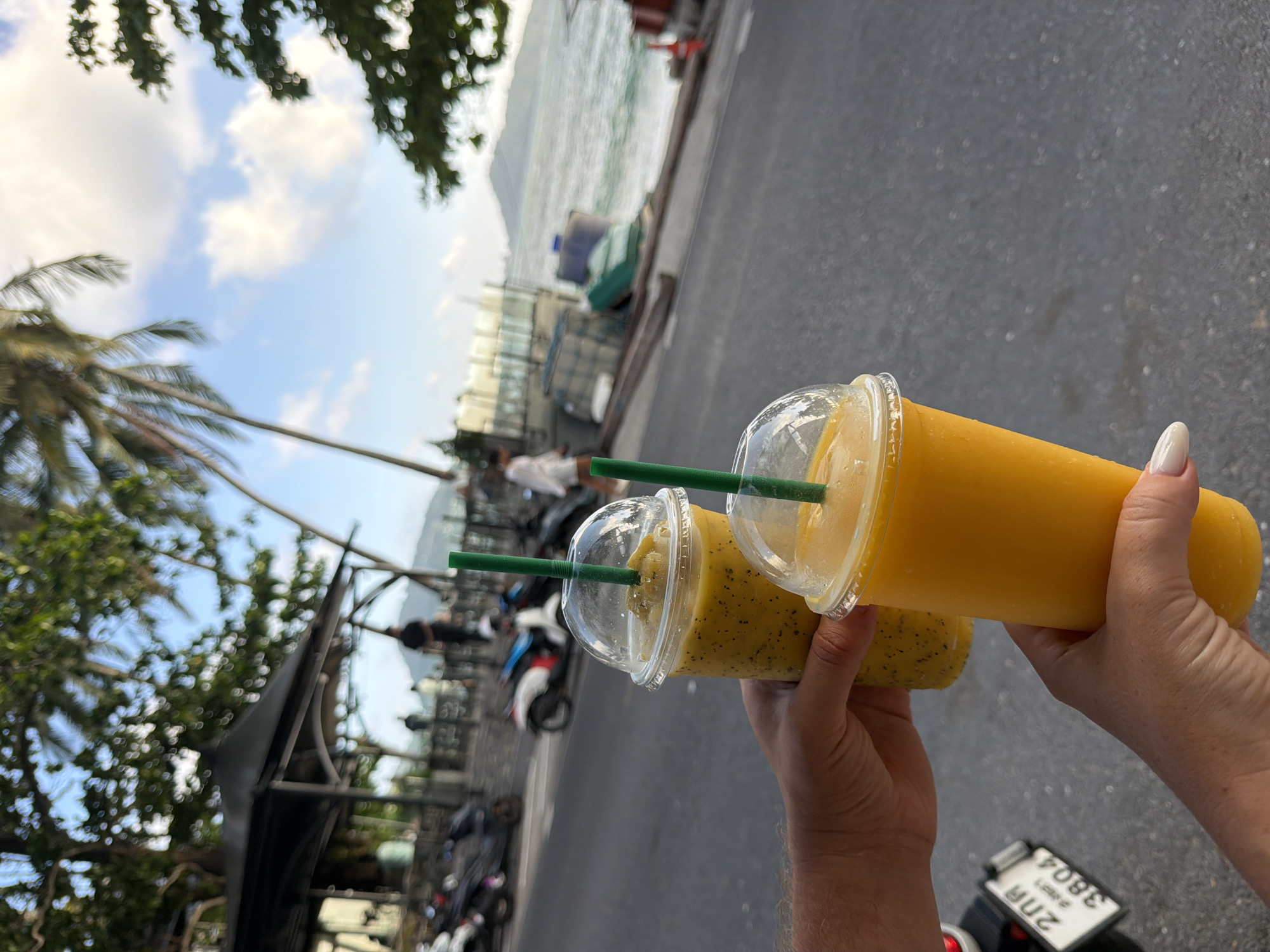 In Patong, Thailand, two hands hold up two large plastic cups filled with yellow smoothies, each topped with a clear plastic lid and a green straw. The smoothie in the foreground is a vibrant, solid yellow, while the one behind it appears to have small dark seeds or fruit pieces mixed in. The background suggests a lively street scene near the beach. Palm trees with fronds sway against a sky with scattered clouds. Parked scooters are visible on the left side of the image, and a road runs horizontally across the lower half. In the distance, the ocean is visible, with a slight wave action and a hazy shoreline. The overall impression is a warm, tropical atmosphere, likely a popular tourist destination.