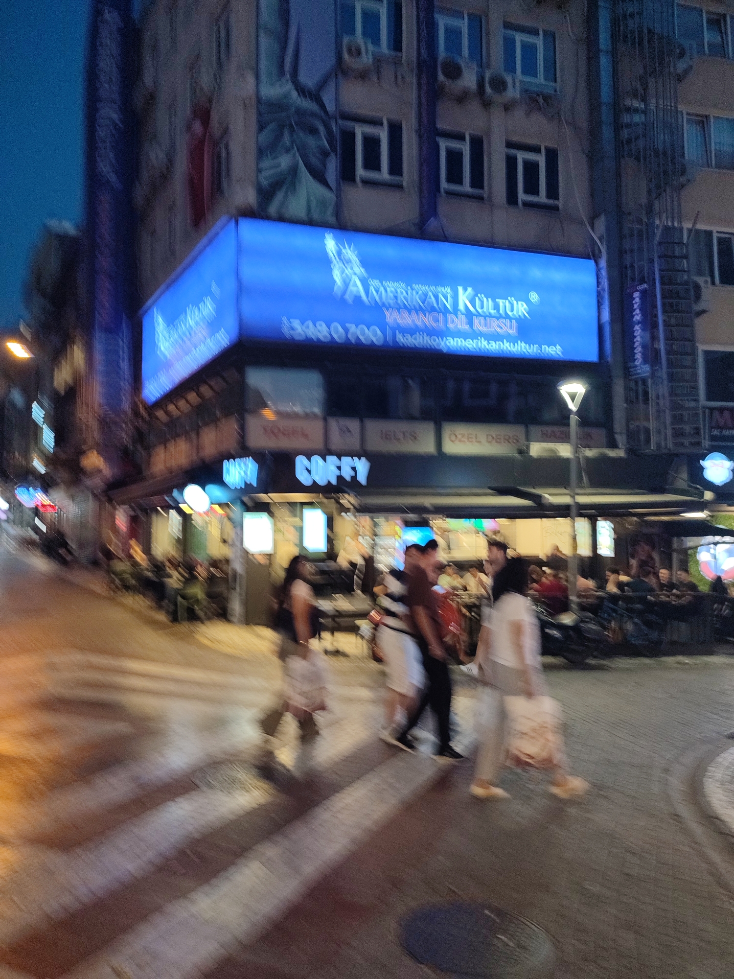 In Kadıköy, Turkey, an evening scene unfolds with a street, a crosswalk, and a building facade. A bright blue illuminated billboard on the building displays the text "AMERİKAN KÜLTÜR YABANCI DİL KURSU" and a website address. Below the billboard, the entrance to a establishment with the name "COFFY" is visible, featuring outdoor seating. Several individuals are captured in motion, walking across the street, some carrying bags. The lighting suggests it is dusk or evening. The overall image is slightly blurred, indicative of movement during the photograph.