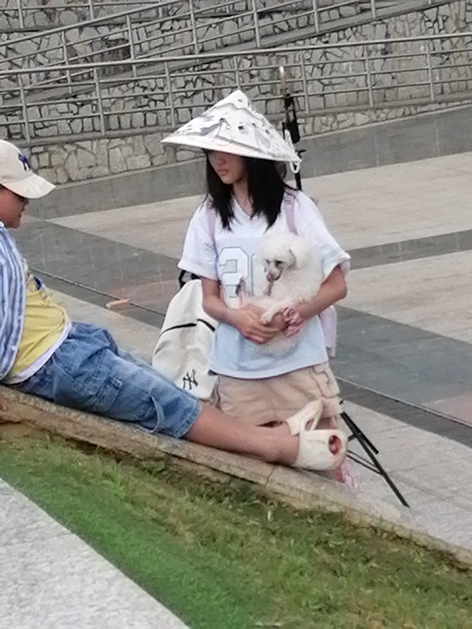 In Da Lat, Vietnam, two individuals are present on an elevated concrete platform with a grassy area below. On the left, a person wearing a blue and white striped shirt, a yellow t-shirt, and blue denim shorts is seated. To the right, a person in a white t-shirt and tan shorts, adorned with a conical hat, is holding a small white dog. The background features a stone wall with metal railings.