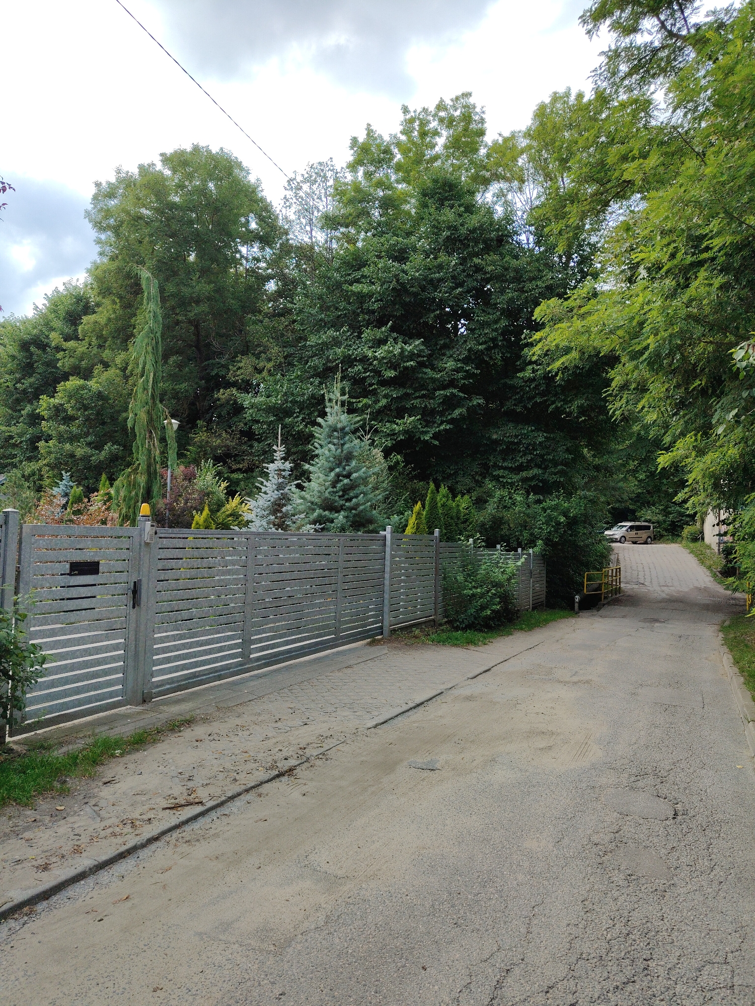 The image depicts a residential driveway in Rumia, Poland, bordered by a grey, horizontal-slatted metal fence on the left and lush green trees on the right. The driveway is paved with asphalt that shows signs of wear and tear, including cracks and uneven patches. At the far end of the driveway, a beige van is parked. A yellow metal railing is visible on the right side of the driveway, next to a small structure. The sky above is overcast with grey clouds. The fence is equipped with a yellow warning light on its leftmost gatepost and a black mailbox. Various evergreen and deciduous trees and bushes are planted behind the fence, creating a densely vegetated backdrop. The ground alongside the driveway is covered in dirt and scattered leaves.
