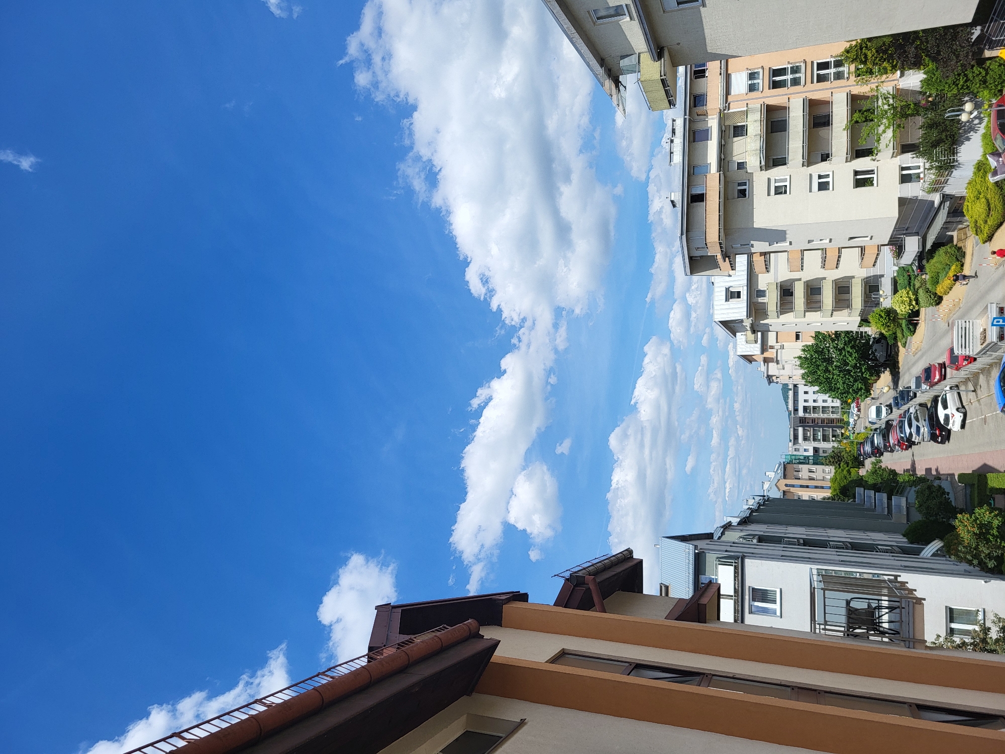 The image shows a street scene in an urban environment. On the right, there are tall apartment buildings with balconies and green foliage. A street runs alongside the buildings, with parked cars of various colors lining the side. A few pedestrians can be seen walking on the sidewalk. The sky takes up a large portion of the left side of the image, showcasing a clear blue sky with scattered white clouds. In the foreground, parts of rooftops and building facades are visible. The architecture suggests a European city, but the specific location is not identifiable from the image alone.