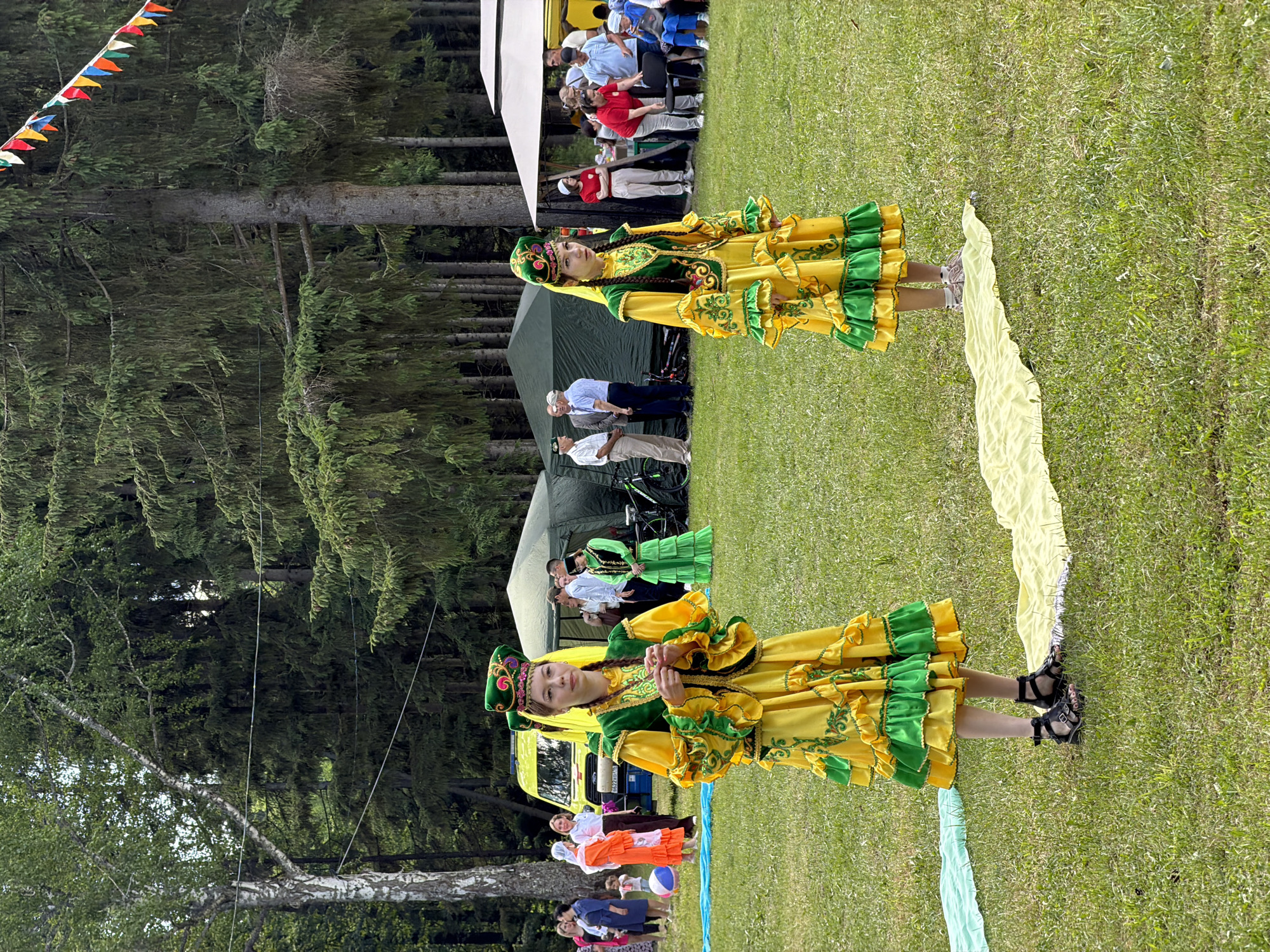 The image captures an outdoor gathering on a sunny day in a grassy, wooded area. Two young women, appearing to be teenagers or young adults, stand in the foreground on a manicured lawn. Both are dressed in elaborate traditional attire featuring vibrant yellow long-sleeved dresses with tiered green ruffles and trim, adorned with intricate gold-colored embroidery. They wear matching green hats with yellow accents, and the woman on the left also has yellow fabric draped over her braided hair. The woman on the left is positioned slightly angled towards the viewer's right, with her hands clasped in front of her. The woman on the right stands facing forward. On the grass directly in front of them are two narrow strips of fabric: one light blue/turquoise to the left and one light yellow/cream extending from the center towards the right woman.
In the midground, behind the two central figures, various activities and people are visible. To the left, a bright yellow van, identified as an emergency vehicle with blue lights on its roof, faces towards the right. Behind it stands a dark green tent. A woman in a similar green traditional dress is visible near this tent, interacting with other individuals dressed in casual attire. Further to the right, a white tent or canopy is set up, under which several people are gathered, some seated and others standing. A person in a red shirt is bending over near a chair, and two bicycles are parked near the dark green tent on the far right.
The background consists of a dense forest composed primarily of tall, green coniferous trees, with a large deciduous tree with white bark visible on the far left. A string of colorful triangular flags (bunting) stretches across the upper right portion of the image, strung between the trees. Several thin wires traverse horizontally through the mid-air within the tree line. The ground throughout the scene is covered in green grass. The lighting suggests daytime.
Location: Unknown, Unknown.