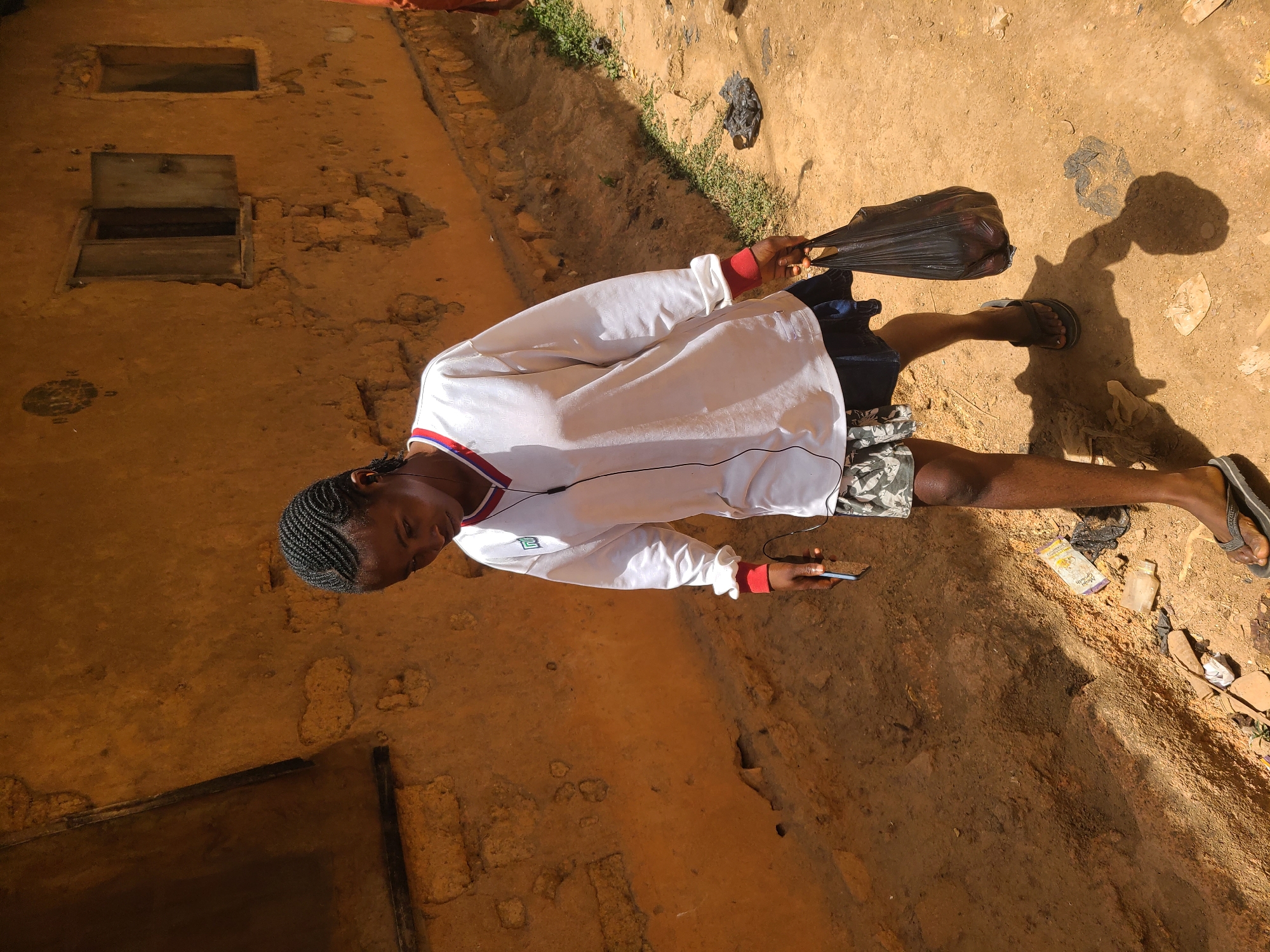 A young, slender-built person stands outdoors in Jos, Nigeria, in an area with dirt ground, amidst scattered debris and trash. The person wears a long-sleeved white shirt with red trim on the collar and sleeves, patterned shorts, and black sandals. They are holding a black plastic bag in one hand and a mobile phone in the other. The person has dark skin and their hair is styled in cornrows. Behind the person is a weathered, yellow-orange colored building or wall with apparent aged cracks and openings. There is also a scattering of trash and various small objects scattered on the ground around the person.