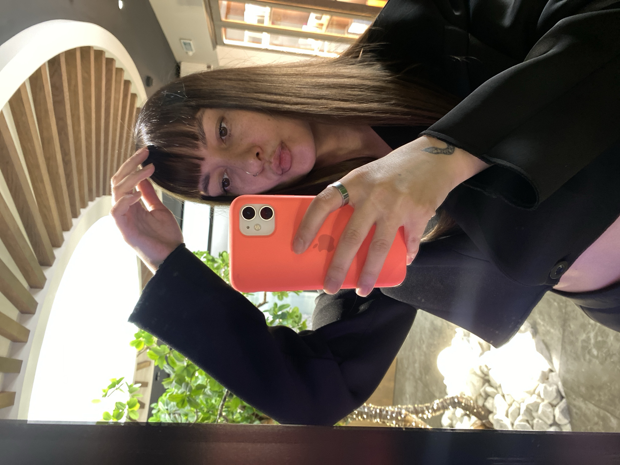 A young woman takes a selfie indoors. She holds a coral-colored iPhone in her right hand, which is adorned with a simple silver ring on her ring finger and a tattoo on her wrist. She has brown hair with bangs and is wearing a black blazer. The background features an arched wooden design, possibly an architectural detail, and plants are visible near a window. The setting appears to be a modernly decorated interior space. There is no information available to establish the city of Votkinsk, Russia as the location.