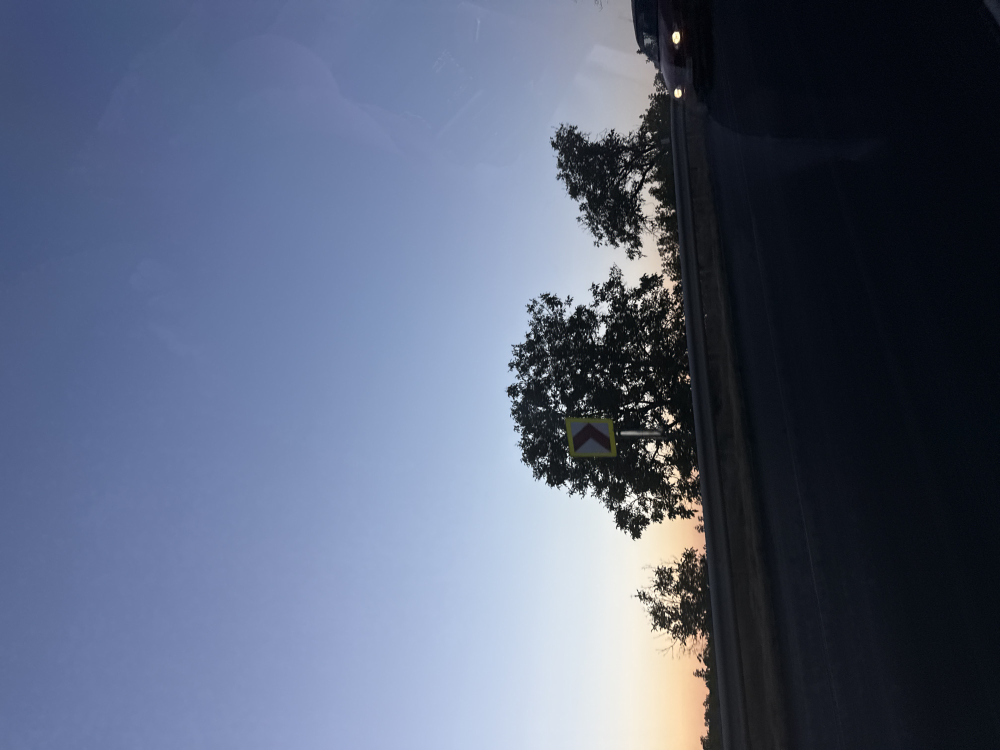 A dusk scene on a highway in Fundul Galbenei, Moldova, is captured from a moving vehicle. The sky transitions from a pale orange on the horizon to a deep blue overhead. Silhouetted trees line the roadside, with a traffic sign indicating a sharp curve to the right. A red car is visible approaching from the right, its headlights illuminated. The road itself is dark, with a reflective barrier separating it from the embankment. The overall impression is one of a journey through a rural landscape as daylight fades.