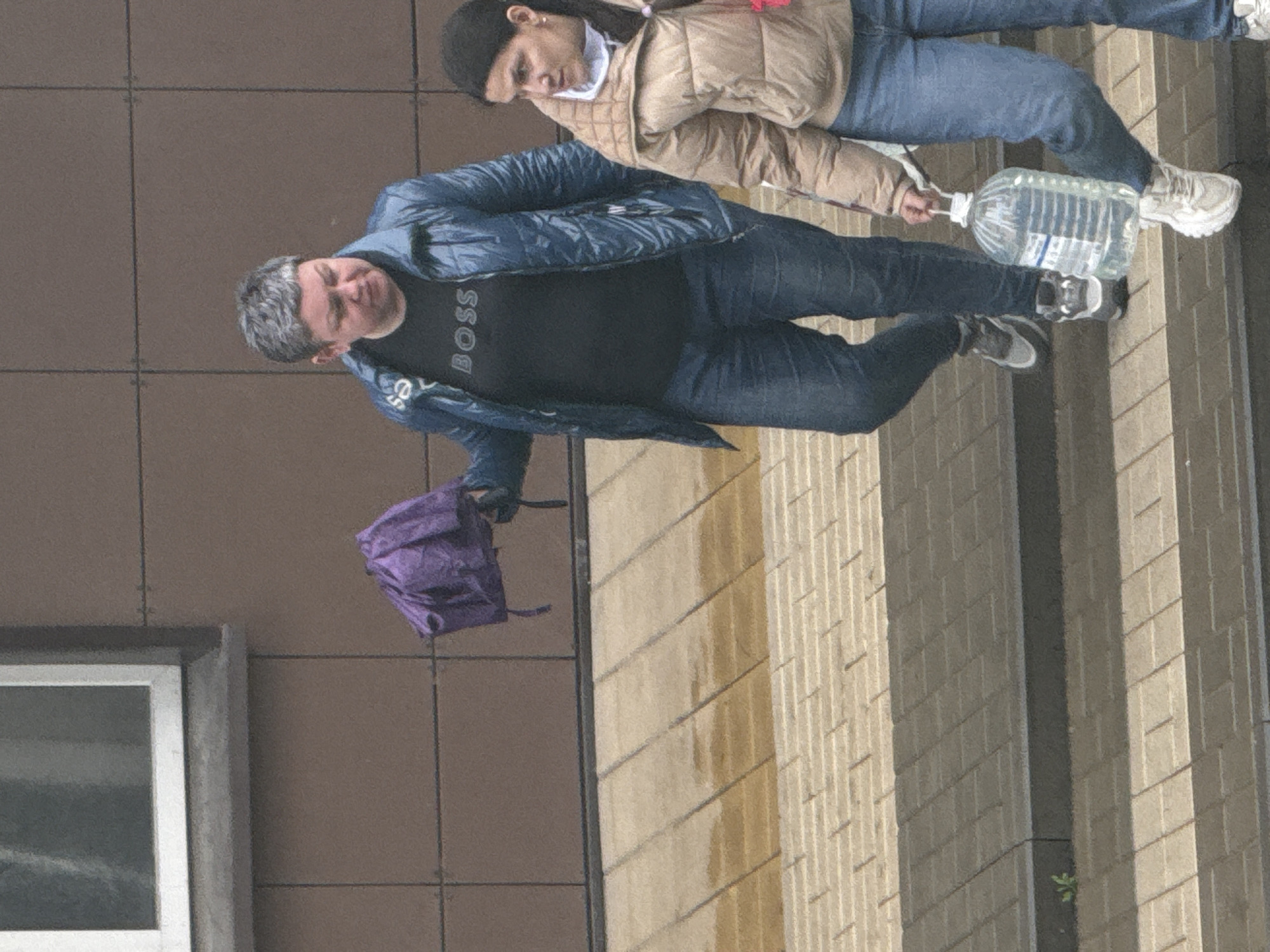 Two individuals are walking down a street in Rostov-on-Don, Russia. The first individual is a man with gray hair, wearing a black "BOSS" t-shirt, blue jeans, and a blue jacket, carrying a purple backpack. The second individual is wearing a beige jacket, blue jeans, and a black head covering. They are jointly carrying a large, clear plastic bottle filled with water. The scene is outdoors, with buildings in the background, and a tiled sidewalk.