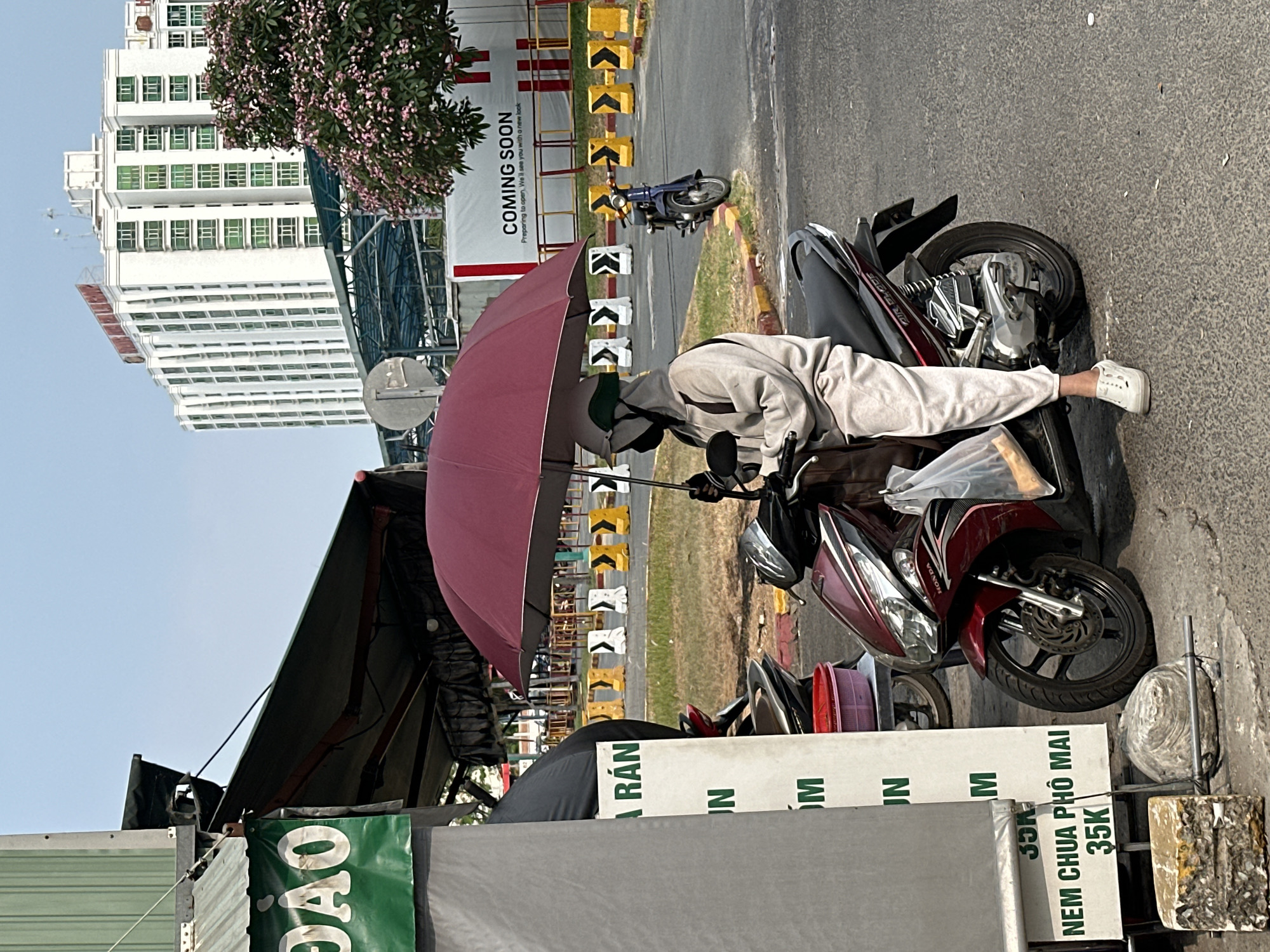 The image captures a street scene in Ho Chi Minh City, Vietnam. A person in light-colored clothing is seated on a red Honda motorcycle, using a maroon umbrella for shade. A food stall is visible on the left, with signs advertising "DAO" and "NEM CHUA PHO MAI 35K". In the background, a building with numerous windows is seen, along with a "COMING SOON" sign and a tree with pink blossoms.