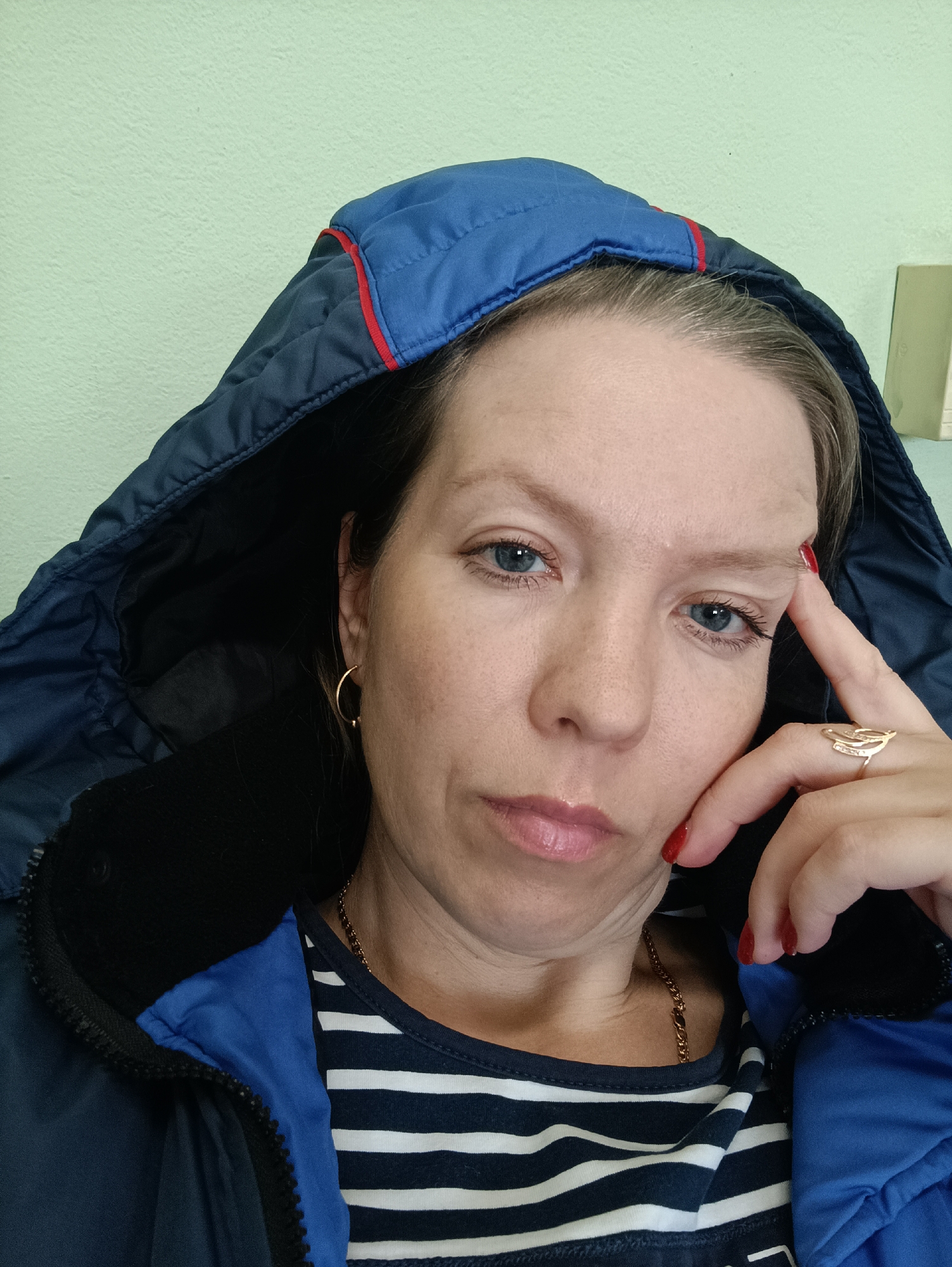 A close-up, indoor portrait features an adult woman, likely in her 30s or 40s, looking directly at the camera. She wears a dark blue hooded jacket with a red trim along the hood's edge, partially obscuring her light brown hair. Beneath the jacket, a navy blue and white striped t-shirt is visible, along with a thin gold chain necklace.
The woman's right hand is raised to her face, with her index finger resting near her temple and her thumb close to her chin, giving her a contemplative or pensive expression. Her fingernails are painted with a glossy red polish. On her right ring finger, she wears a distinctive gold-toned ring. A small gold hoop earring is visible in her left ear. Her eyes are a striking blue, and her lips have a slight pout.
The background is a plain, light green wall, suggesting an indoor setting. The image is brightly lit, but no specific time of day can be inferred. While the jacket might imply cooler weather, the context of Chaikovsky, Russia, is provided externally and not visually indicated within the frame. There is no visible text in the image.