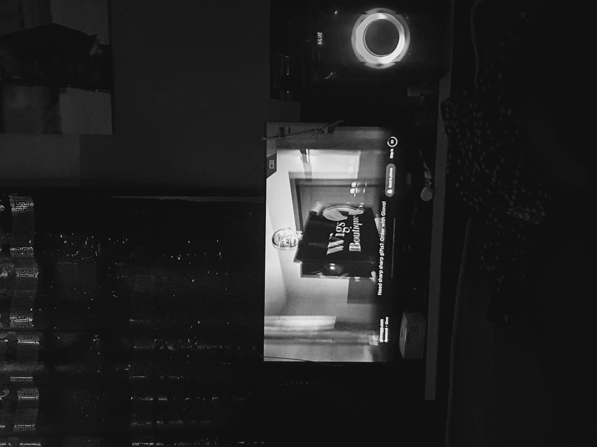The image depicts a television displaying an advertisement for "Wigs Boutique." Text on the screen reads, "Need sharp sharp gifts? Order with Glovo!" The URL "glovoapp.com" is also visible, along with the label "Sponsored • Glovo." A Roku device is located above the TV. The surrounding environment is dark, revealing parts of the wall, furniture, and decorative items. There's no evidence to confirm the location in Lokoja, Nigeria.