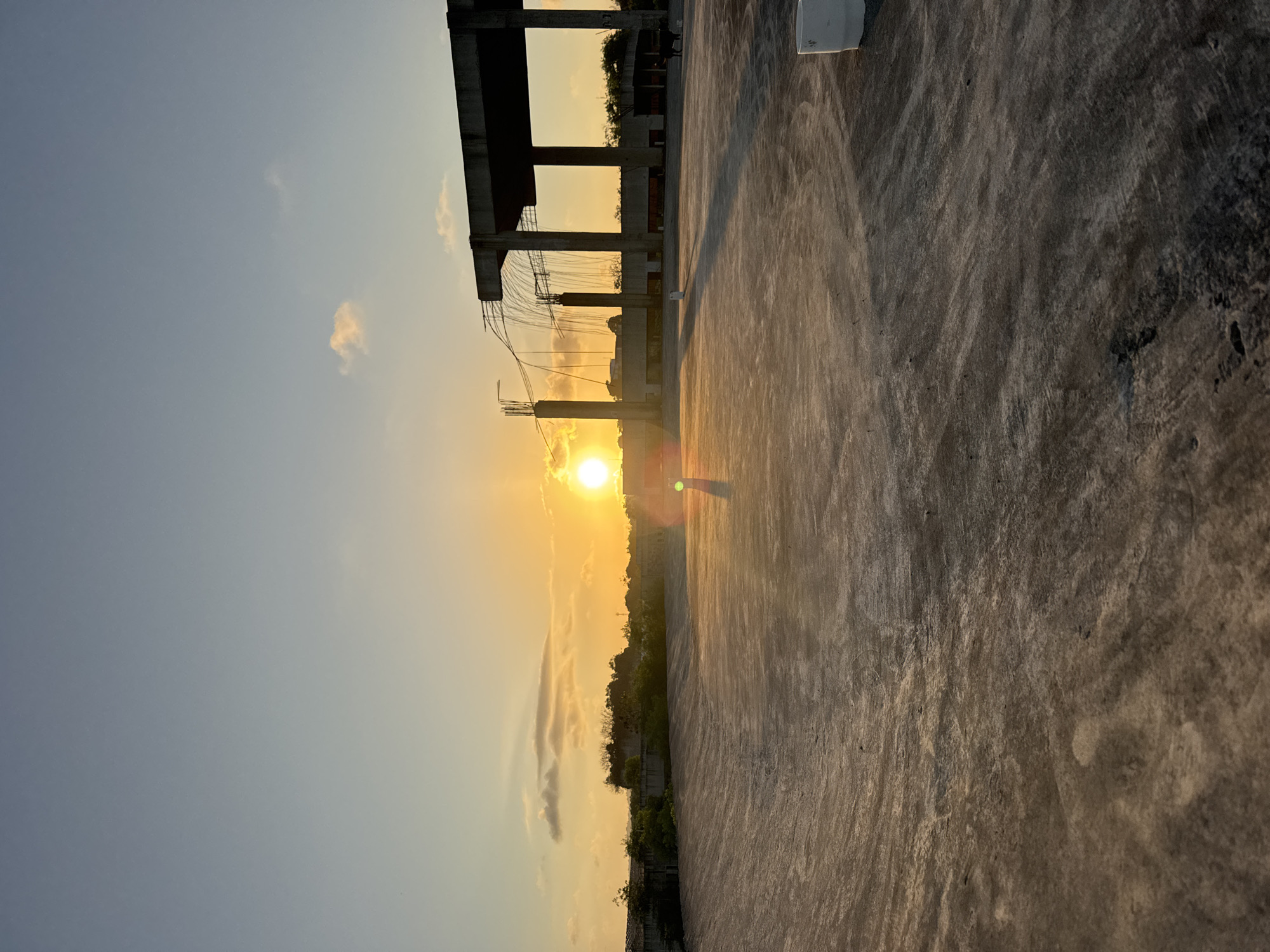The image depicts a rooftop in Kuta Selatan, Indonesia, at sunset. The foreground is dominated by a vast expanse of concrete, showing signs of wear and tear with irregular textures and patterns. A low-lying wall or structure can be seen in the mid-ground, partially obscured by greenery.
Dominating the right side of the mid-ground and background is an unfinished concrete structure, resembling the beginnings of a large building or stadium. It features tall, square pillars supporting a flat, expansive roof. Exposed rebar and dangling wires suggest ongoing construction. The setting sun casts a warm, orange glow across the scene, creating long shadows on the concrete surface.
To the left of the concrete structure, the sun is visible low on the horizon, radiating bright light and creating a lens flare. The sky transitions from a pale blue at the top to a vibrant orange near the horizon, with a few scattered clouds tinged with the sunset colors.
In the far background, a line of trees and foliage is visible, with some buildings peeking through. On the right side of the unfinished structure, a dark-colored quadruped, possibly a dog or a small cow, is standing. Two white cylindrical objects, likely buckets or containers, are placed on the rooftop, one in the mid-ground and another in the lower right corner. The overall impression is one of an industrial or construction site bathed in the warm light of a tropical sunset.