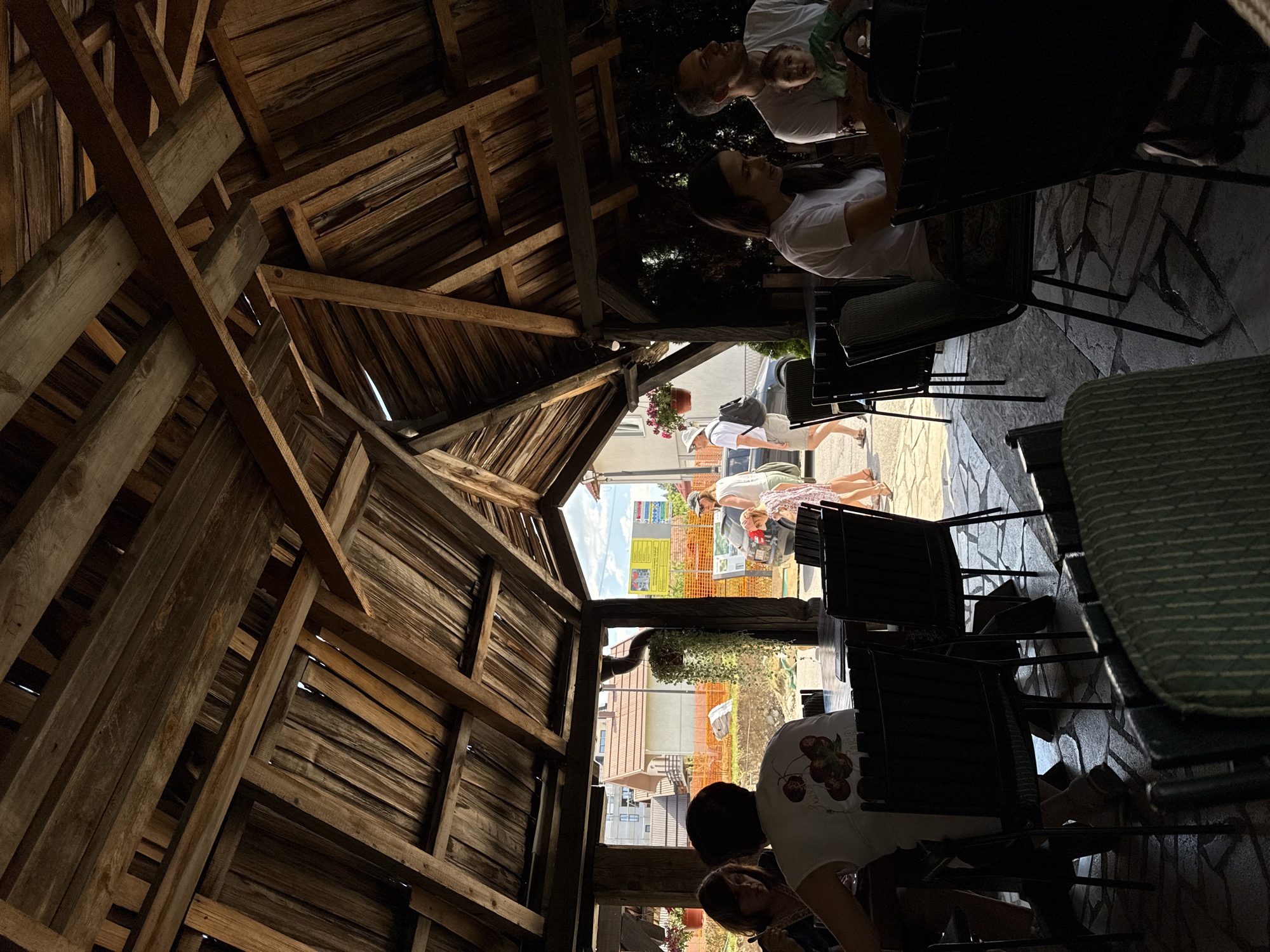 The image captures a lively outdoor seating area of a restaurant in Kolašin, Montenegro, on a sunny day. The open-air structure, constructed from rustic wooden beams and planks, provides shade and a natural ambiance. Several people are visible enjoying the setting: a woman and a man sit at a table on the right, the man holding a baby. In the center, a woman in a white shirt with a cherry design sits at a table, facing away from the camera. Another group of people, including a woman in a pink dress and a man in a white shirt and hat, walk in the background. The ground is paved with natural stone. Beyond the seating area, construction is visible with orange netting and buildings under construction, indicating ongoing development in the city. The scene suggests a casual and social atmosphere, likely a popular spot for dining and relaxation in Kolašin.
