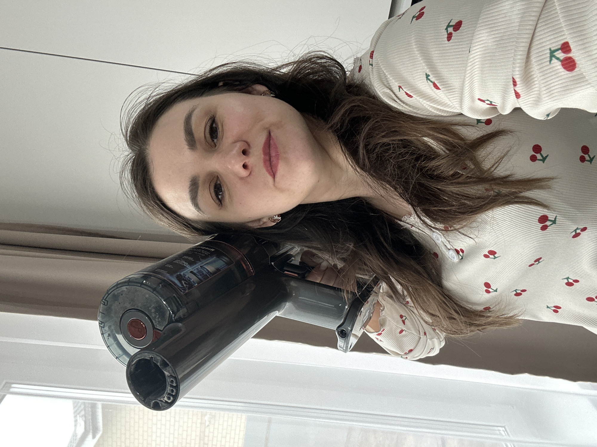 A medium close-up shot captures a young woman indoors, holding a dark gray handheld stick vacuum cleaner. The image is taken at a slight upward angle, with the woman looking directly at the camera. She has long, dark brown hair that falls over her shoulders, dark eyebrows, and is wearing light makeup with pinkish lipstick. Her expression is neutral to slightly pleasant, with a faint smile. She wears a cream or off-white long-sleeved top with a ribbed texture, patterned with small red cherries and green stems. Small, possibly gold-toned, earrings are visible.
The vacuum cleaner, held by the woman, is prominent in the foreground. It has a transparent dustbin section, a red circular button or indicator, and a wide nozzle at the end. Some indiscernible text is visible on the side of the vacuum's body.
Behind the woman, a bright window is visible, letting in natural light and suggesting it is daytime. Outside the window, faint brickwork is discernible, indicating another building or structure. A white window frame or sill is also part of the background. The scene depicts a casual, domestic moment in an indoor setting.
