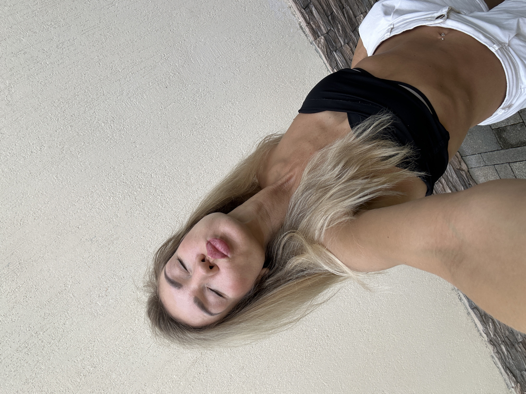A blonde woman with her eyes closed and lips pursed is posing for a selfie against a textured beige wall. She is wearing a black sports bra and white shorts, with a silver belly button piercing visible. The lower part of the image shows a stone-tiled ground.