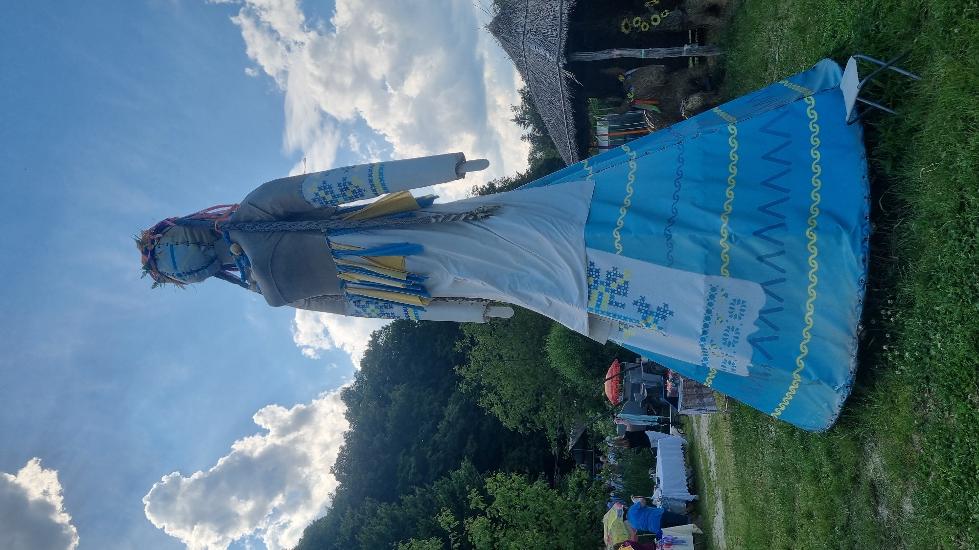 This image, taken in Melnyky, Ukraine, captures an outdoor scene dominated by a monumental anthropomorphic figure, likely a doll or effigy, standing prominently in a grassy field under a bright blue sky with scattered white clouds. The figure, dressed in what appears to be stylized traditional Ukrainian attire, features a light-colored fabric head with stitched seams and a blue cross-like design. It wears a floral and leaf crown with colorful ribbons and braids extending from the head. The torso is a solid gray fabric, while the long white sleeves are adorned with blue and yellow embroidered patterns reminiscent of vyshyvanka designs. The figure's skirt is multi-layered, with an upper white section from which blue and yellow fabric strips hang. The lower skirt expands into a large, conical base, predominantly blue, and decorated with elaborate white, blue, and yellow patterns, including cross-stitch motifs, wavy lines, and chevrons. A small, white foldable stool rests at the base of the figure on the right.
In the mid-ground, to the left, a vibrant outdoor market or fair is visible, with people browsing items on white-covered tables. A person in a blue shirt and yellow hat stands near a table, while another in a black shirt holds up a white textile. An orange umbrella is partially seen among the stalls, and a vehicle is in the background. Dense green trees and foliage form a natural backdrop behind the market activity. To the right of the central figure, a small, rustic hut with a thatched roof is present. Nearby, hay bales shaped like animals, adorned with colorful ribbons and sunflowers, are displayed around a wooden post. The ground consists of green grass with small patches of clover.
