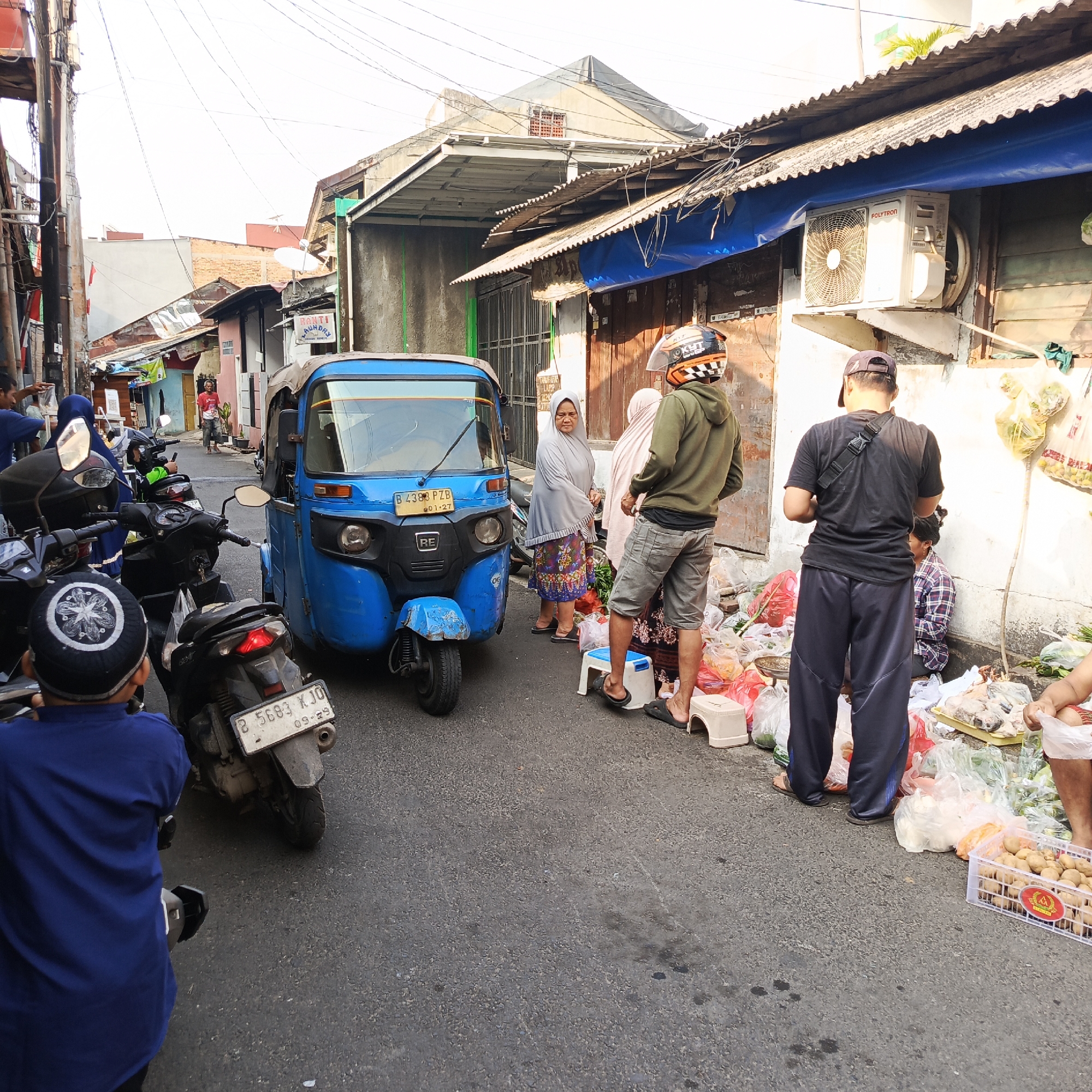 The image depicts a street scene in South Jakarta, Indonesia, likely a market area. A blue three-wheeled vehicle, a bajaj, is parked in the middle of the narrow street. Several motorcycles are parked along the sides. A group of people are gathered, some appearing to be vendors and others customers. One individual, wearing a green hoodie and a helmet, stands near a display of produce. Another person, in a black t-shirt and dark pants, is facing away from the camera. Two women, one in a grey hijab and patterned skirt, and another in a pale pink hijab, are also present. The surrounding buildings appear to be made of corrugated metal and concrete, with some displaying air conditioning units and electrical wiring. Bags of produce, including potatoes and leafy vegetables, are visible on the ground and in crates, suggesting a marketplace. The time of day appears to be daytime, with bright sunlight casting shadows.