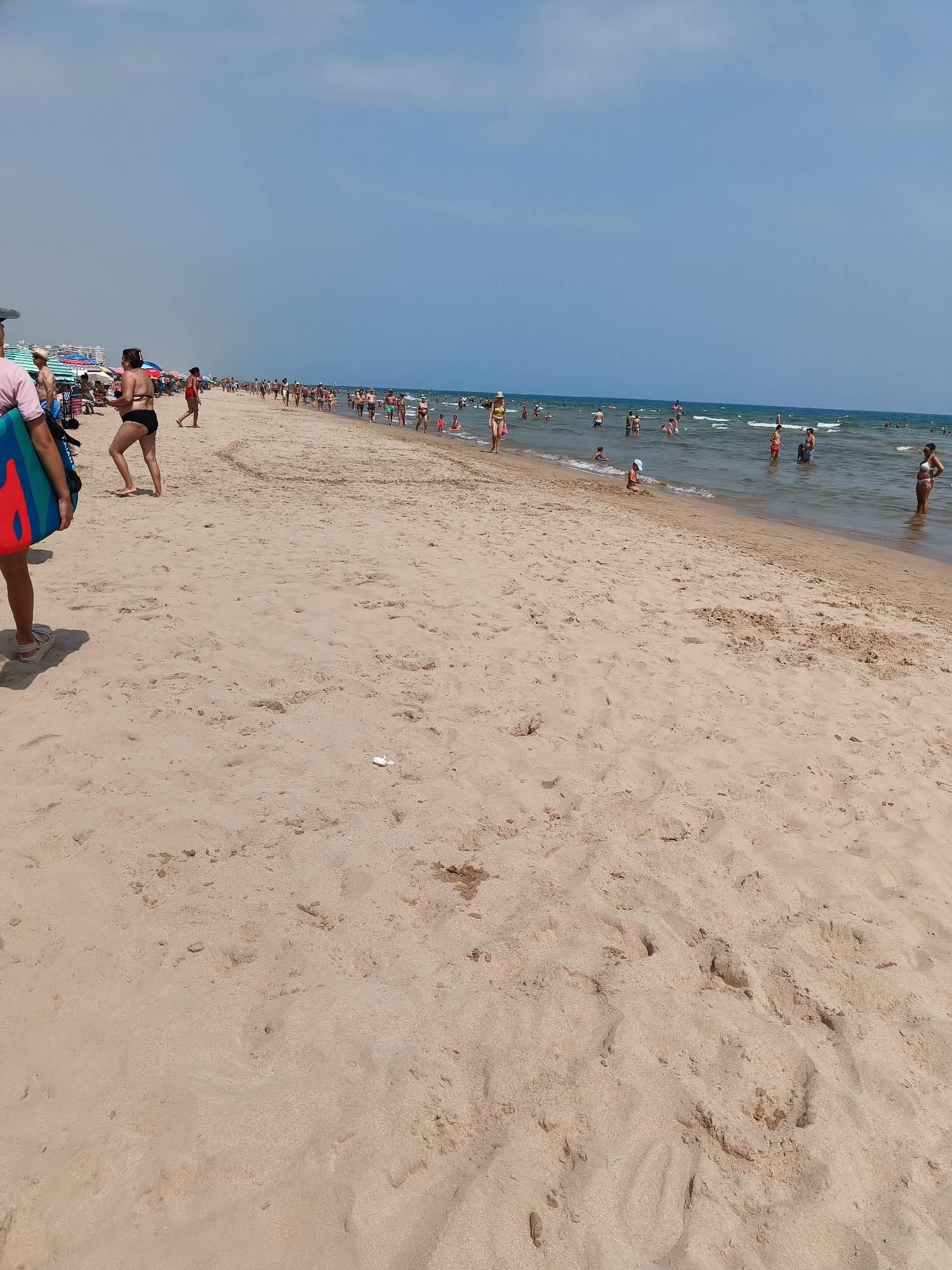 The image depicts a sunny day at Grau I Platja, Spain, a beach populated by numerous individuals. The foreground is dominated by a vast expanse of light-colored sand, marked by footprints and some scattered debris. In the middle ground, a diverse crowd is engaged in various beach activities. Some people are wading in the shallow, calm water, while others are walking along the shoreline or standing in groups. Further back, more people are visible in the water, some swimming and others gathered closer to the shore. The background shows a clear blue sky with a few wispy clouds, and the gentle waves of the sea stretching towards the horizon. A line of umbrellas and beach chairs is discernible on the sand near the water's edge, suggesting a developed beach area. A person in the left foreground is partially visible, carrying a bodyboard. The overall scene conveys a lively and typical summer beach atmosphere.