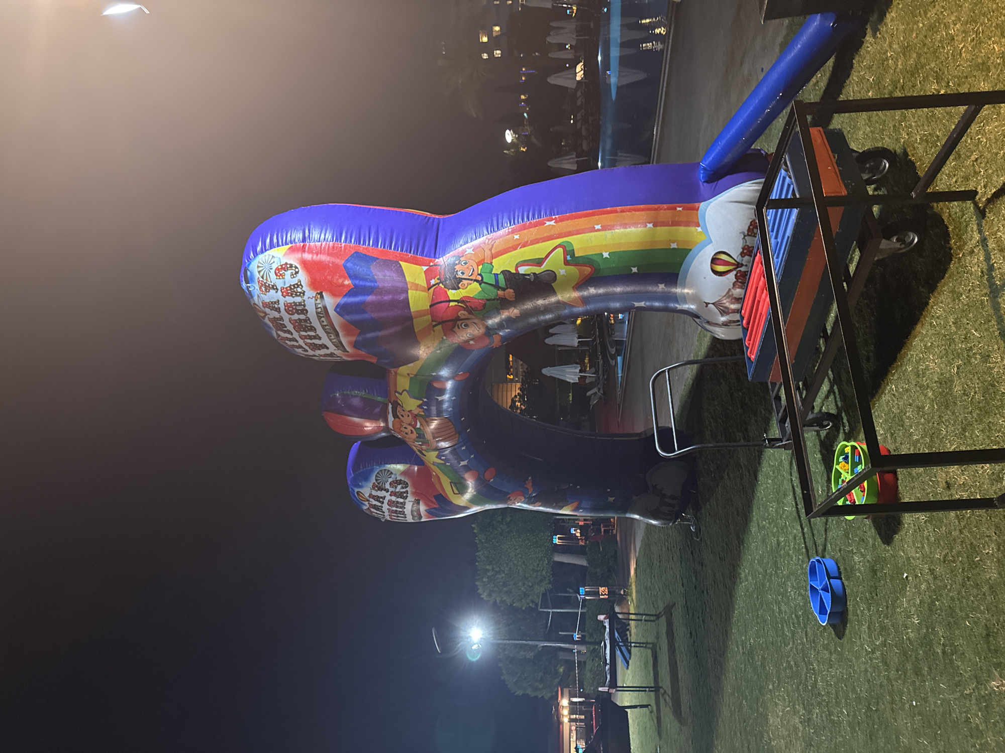 An illuminated, inflatable archway with circus-themed artwork and the words "FAMILY CARNIVAL" is set up on a grassy area at night in Aksu, Turkey. The archway features colorful illustrations of children, a hot air balloon, and a circus tent. In the foreground, a metal cart with red and blue cylindrical objects is parked on the grass. Nearby, a blue plastic organizer and a green and red container with colorful plastic blocks are visible. In the background, a swimming pool with illuminated water and lounge chairs is present, along with trees and buildings. The scene appears to be an outdoor event or entertainment area.