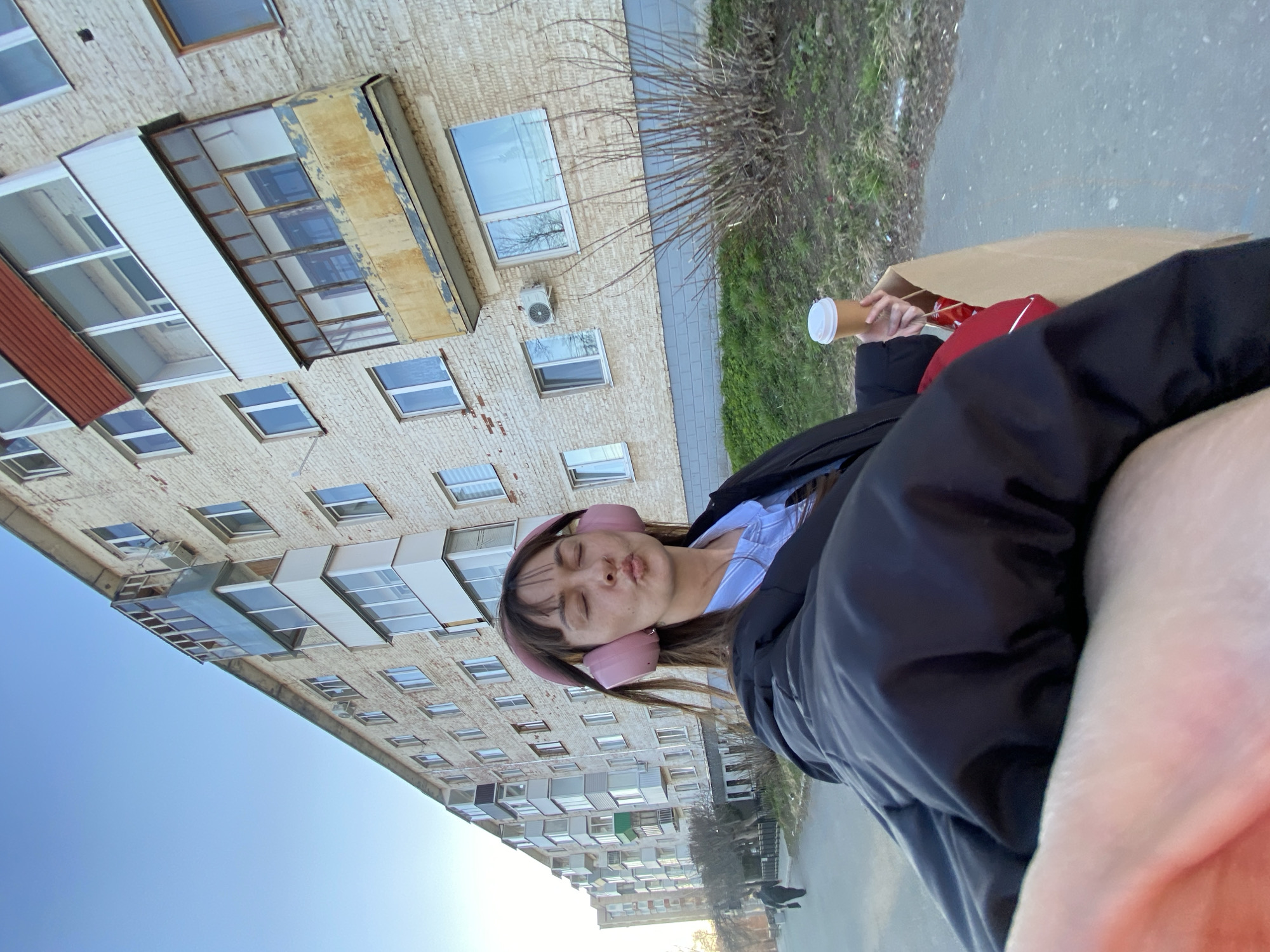 A medium shot shows a person outdoors. The person is fair-skinned and has dark hair and pink headphones. They are wearing a dark jacket and a light-colored shirt. The person is holding a paper bag and a coffee cup. In the background is a multi-story building with light-colored brick and many windows. There is a sidewalk and some grass visible as well. The location may be in Votkinsk, Russia.