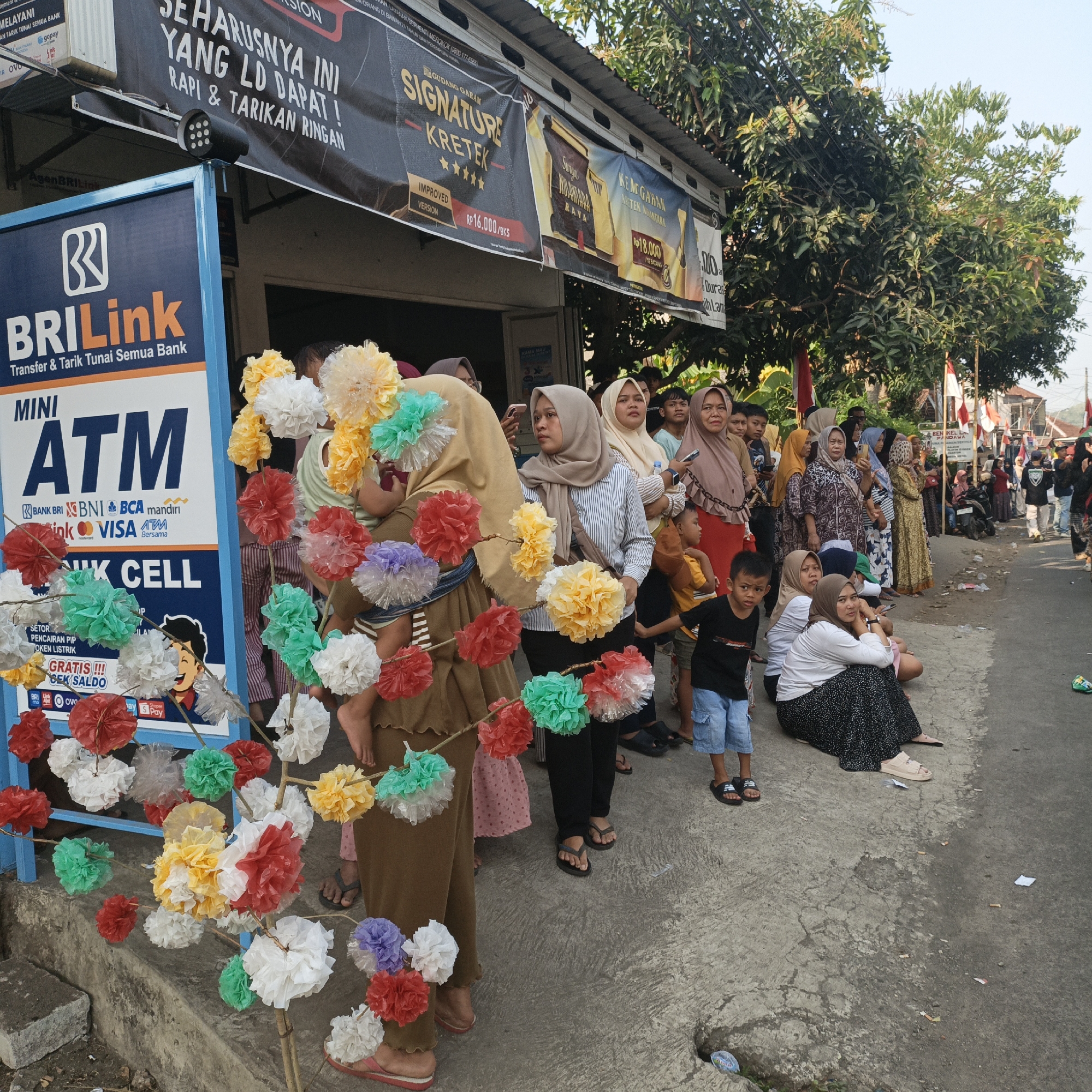 This image depicts a street scene in Tanahbaya, Indonesia, featuring a gathering of people near a BRI Link mini ATM and a shop. The scene is crowded with individuals of various ages, many of whom are women wearing headscarves, indicating a likely Indonesian cultural context. In the foreground, colorful paper pom-poms are displayed on sticks, some held by a woman in a brown tunic. A BRI Link sign for an ATM is prominently displayed on the left.
Behind the ATM, a shop is visible with advertising banners, one of which appears to be for Gudang Garam cigarettes. The banners are written in Indonesian. Several people are standing and sitting on the side of the road, some looking towards the camera, while others are engaged in conversation. The environment appears to be an urban or semi-urban setting, with buildings, trees, and a paved road. Indonesian flags are visible in the background, suggesting a possible national celebration or event. The ground is concrete and appears to be somewhat littered with small pieces of paper and what look like cigarette butts. The lighting suggests it is daytime.