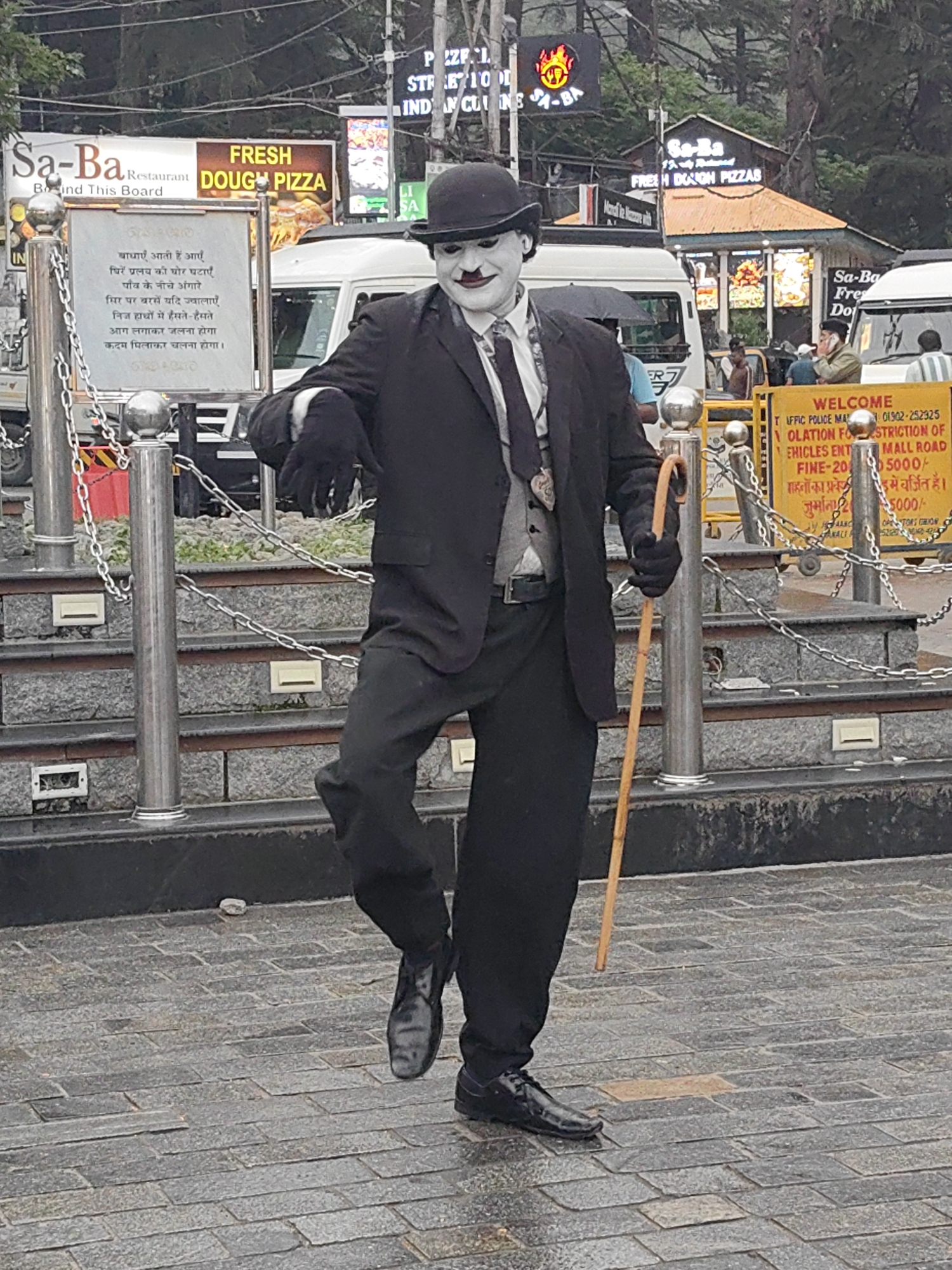 The image depicts a street performer dressed as Charlie Chaplin in Manali, India. The performer is wearing a black suit, bowler hat, white face paint with dark makeup around the eyes and mouth, and a tie. They are holding a cane and are positioned on a paved area with stone steps and metal railings in the background. Signs for "Sa-Ba Restaurant" and "Fresh Dough Pizza" are visible behind the performer, indicating a commercial area. The overall scene suggests a public space, likely a tourist area, where the performer is engaging with onlookers.