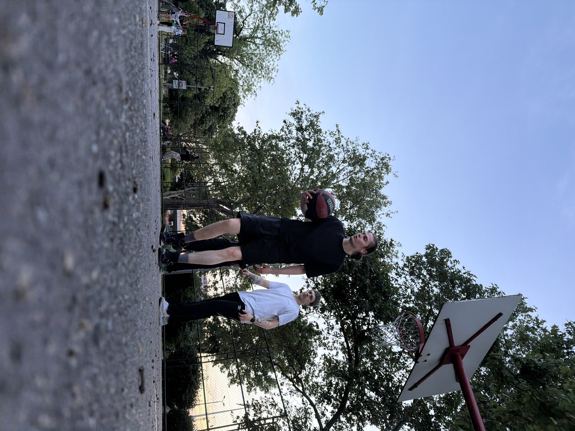 Two men stand on a basketball court. One holds a basketball; both appear to be on the court to play. The basketball court is adjacent to a metal fence. Beyond the fence, trees and green foliage are visible, suggesting a park or green space. The photo shows a basketball hoop and backboard. Other people are in the background further up the court. The location may be Belgrade, Serbia.