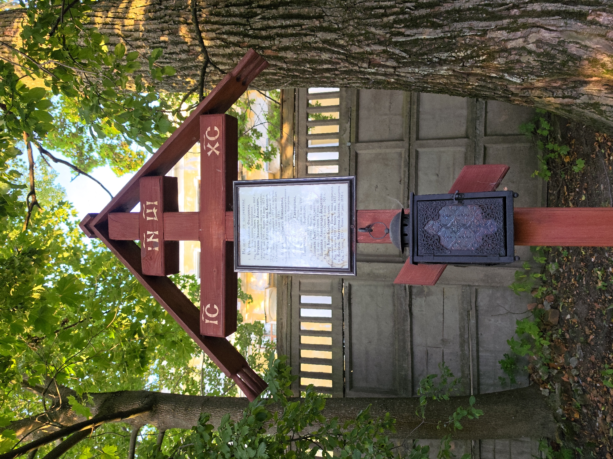 The image shows a wooden Orthodox Christian cross situated in Strelna, Russia. The cross is painted red and features Cyrillic inscriptions. Attached to the front of the cross is a dark metal lantern with decorative filigree and colored glass panels. A framed document with text in Cyrillic is also affixed to the cross. The cross is positioned outdoors, with a gray concrete fence visible behind it. Two large trees with leafy canopies are present on either side of the cross, with sunlight filtering through the leaves. The ground beneath the cross is covered in dirt, leaves, and some debris. The overall scene suggests a memorial or religious marker in a natural or park-like setting.