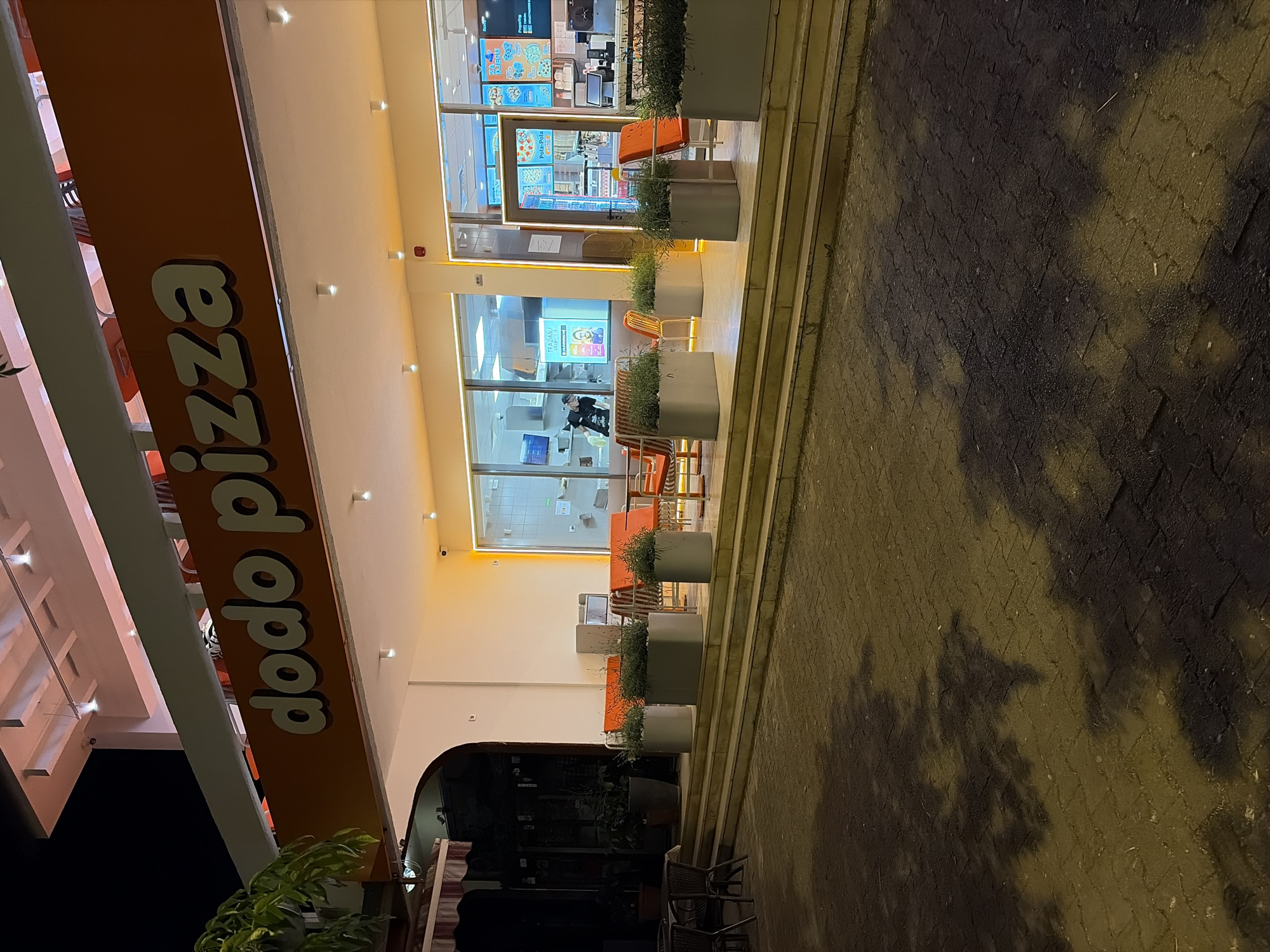 The image depicts the exterior of a Dodo Pizza restaurant at night. The sign "dodo pizza" is prominently displayed on the building. The restaurant has large windows, and seating areas are visible inside and outside. There are planters with greenery along the front of the restaurant. The sidewalk outside the restaurant is paved. The location is the city of Bar, Montenegro.