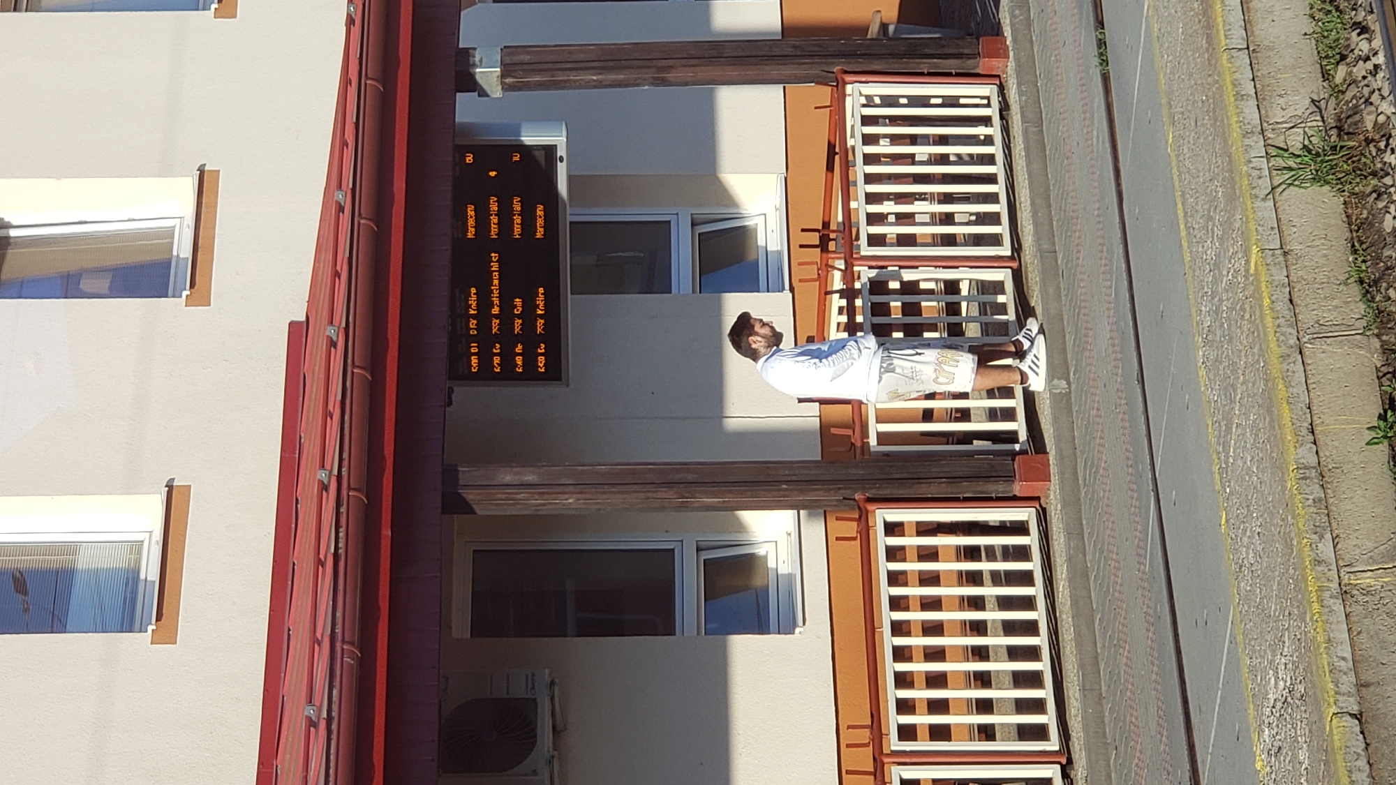 The image shows a man standing on a train platform in Spišská Nová Ves, Slovakia, looking down at his phone. He is wearing a white long-sleeved shirt and white shorts with a pattern on them, and white Adidas sneakers. Behind him, a digital display shows train departure times and destinations. The platform is covered by a red awning, and there is a white railing separating the platform from the tracks. The building behind the platform is light-colored with windows. The overall scene appears to be a sunny day at a train station.
