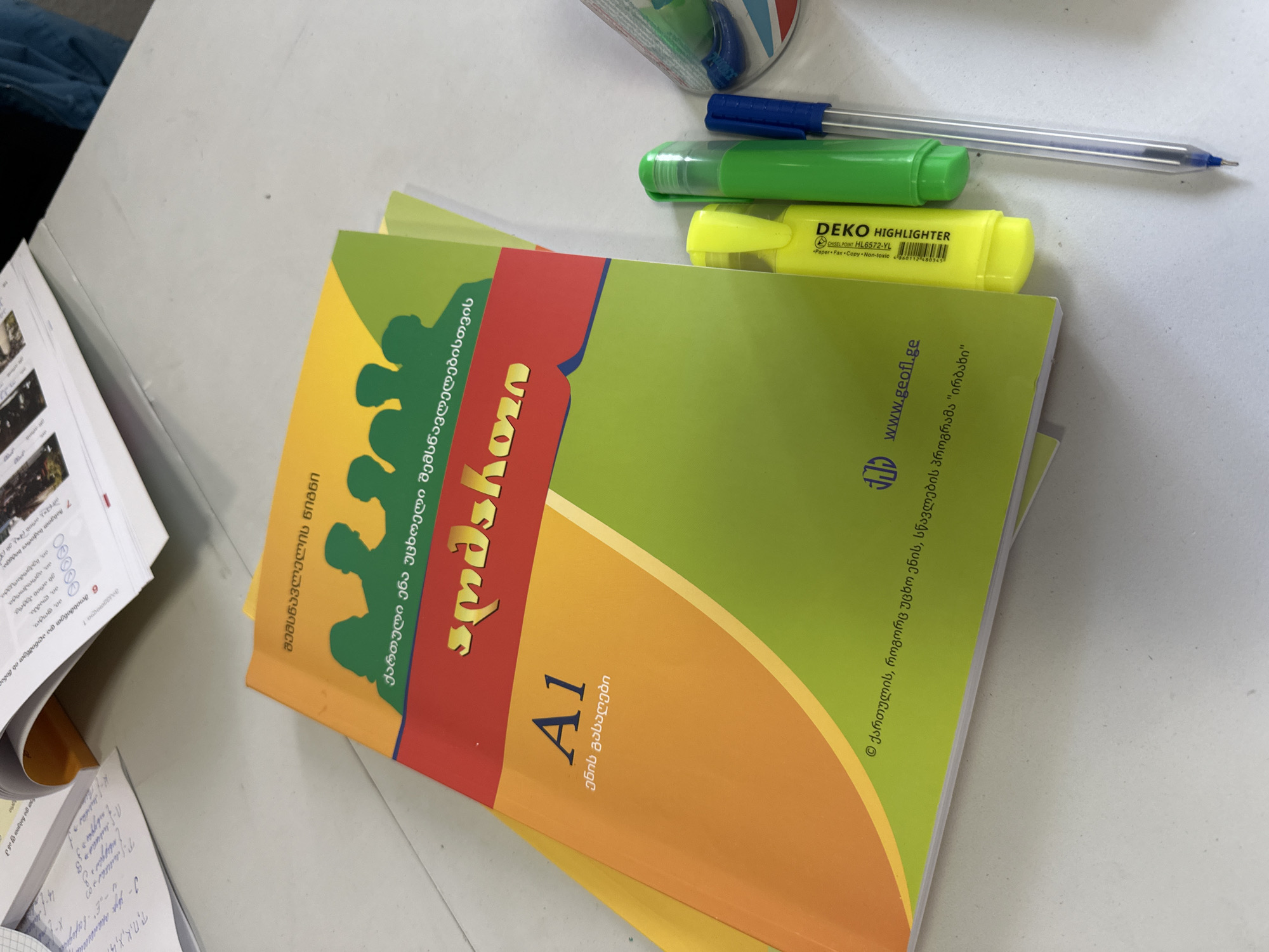 The image captures a study scene on a light-colored desk, featuring an open Georgian language textbook, a notebook, and writing utensils. The prominent object is a vibrant textbook for "Georgian language for foreigners," titled "სმარტჰაუსი" (Smart House), designated for "A1" proficiency level, and identified as a "Learner's Book" and "Language Key." Its cover also displays "www.geofl.ge" and a copyright for the "Irbakhi" Georgian language teaching program.
Next to the textbook, an open notebook reveals handwritten Georgian text and numbers, along with a printed section titled "Lesson 1" that includes the phrase "ეს არის პეტრე." (This is Peter.) and instructions like "Listen and mark the..."
A blue pen, a green marker, and a yellow DEKO HIGHLIGHTER (model HL6572-YL, chisel point, non-toxic, with barcode 4860112480345) are placed on the desk, suggesting an active study session. The setting is indoors, without specific indicators of time or weather. The overall activity depicted is language learning or desk work, likely in a classroom or personal study environment in Tbilisi, Georgia, given the context of the Georgian language materials.
